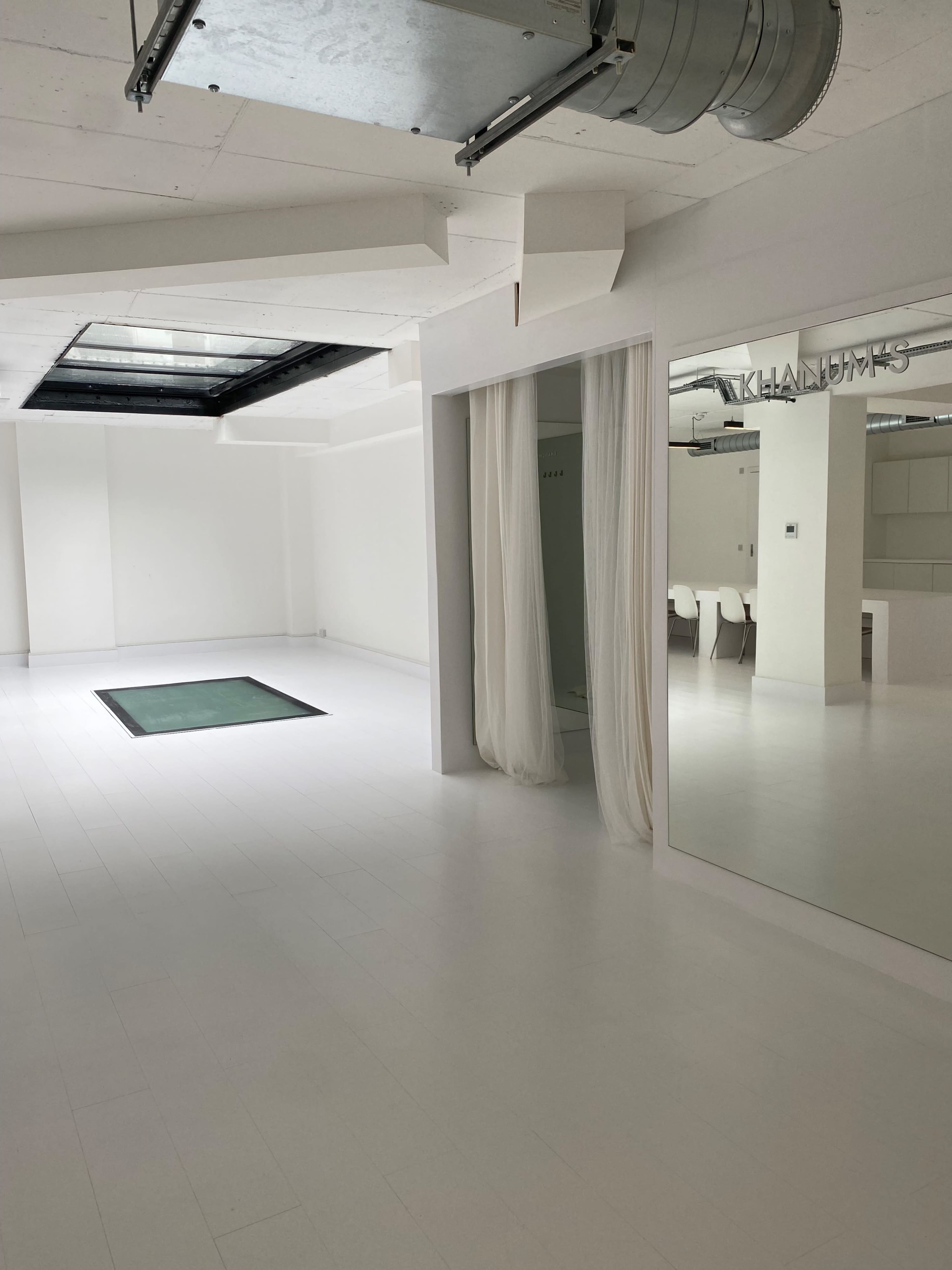 A spacious, empty white room with a rectangular area covered in green, sheer drapes on one side, and a large mirror reflecting the interior on the opposite side.