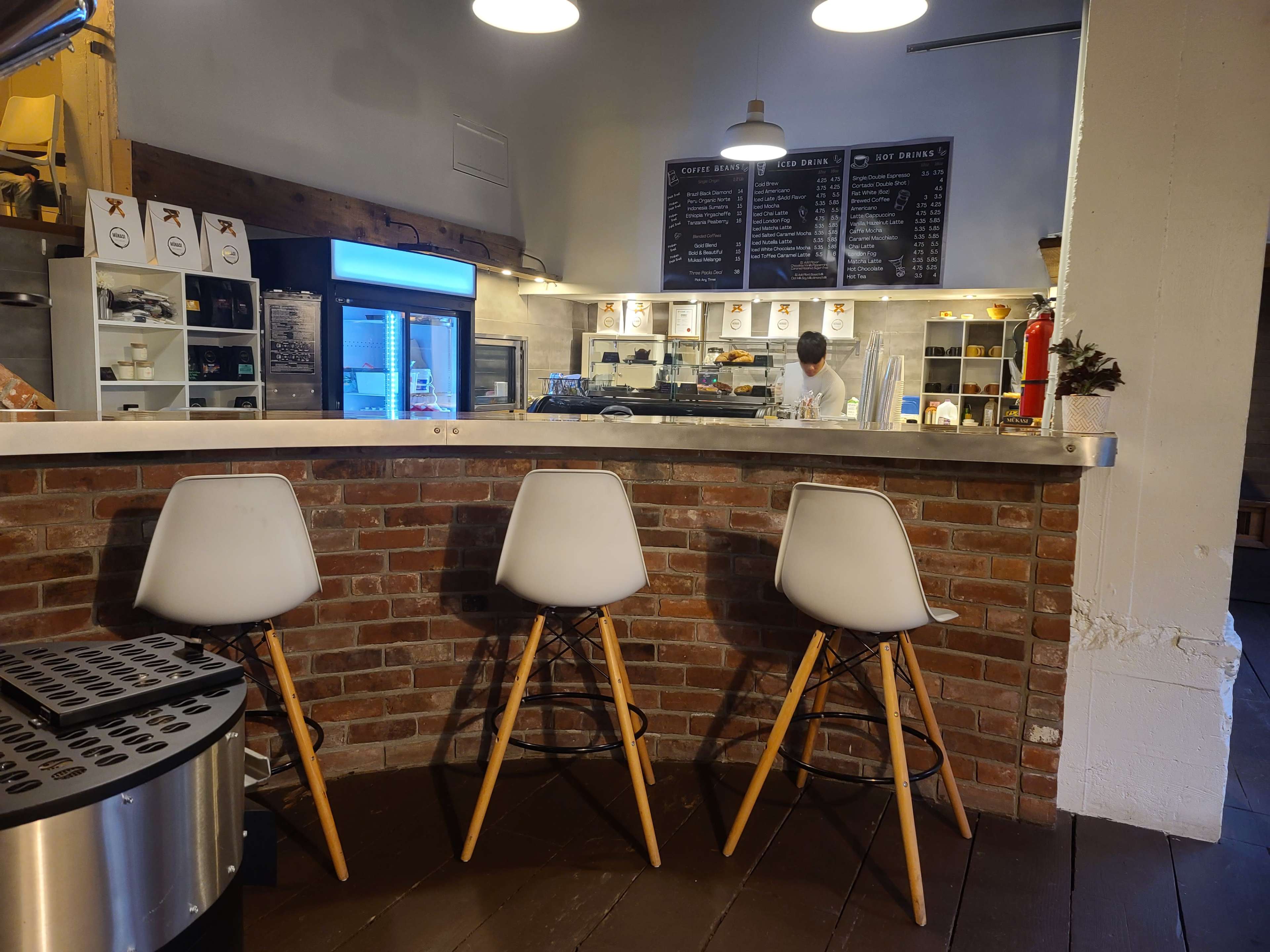 A bar area with three white chairs sits in front of a kitchen space featuring a chef and a menu board on the wall.