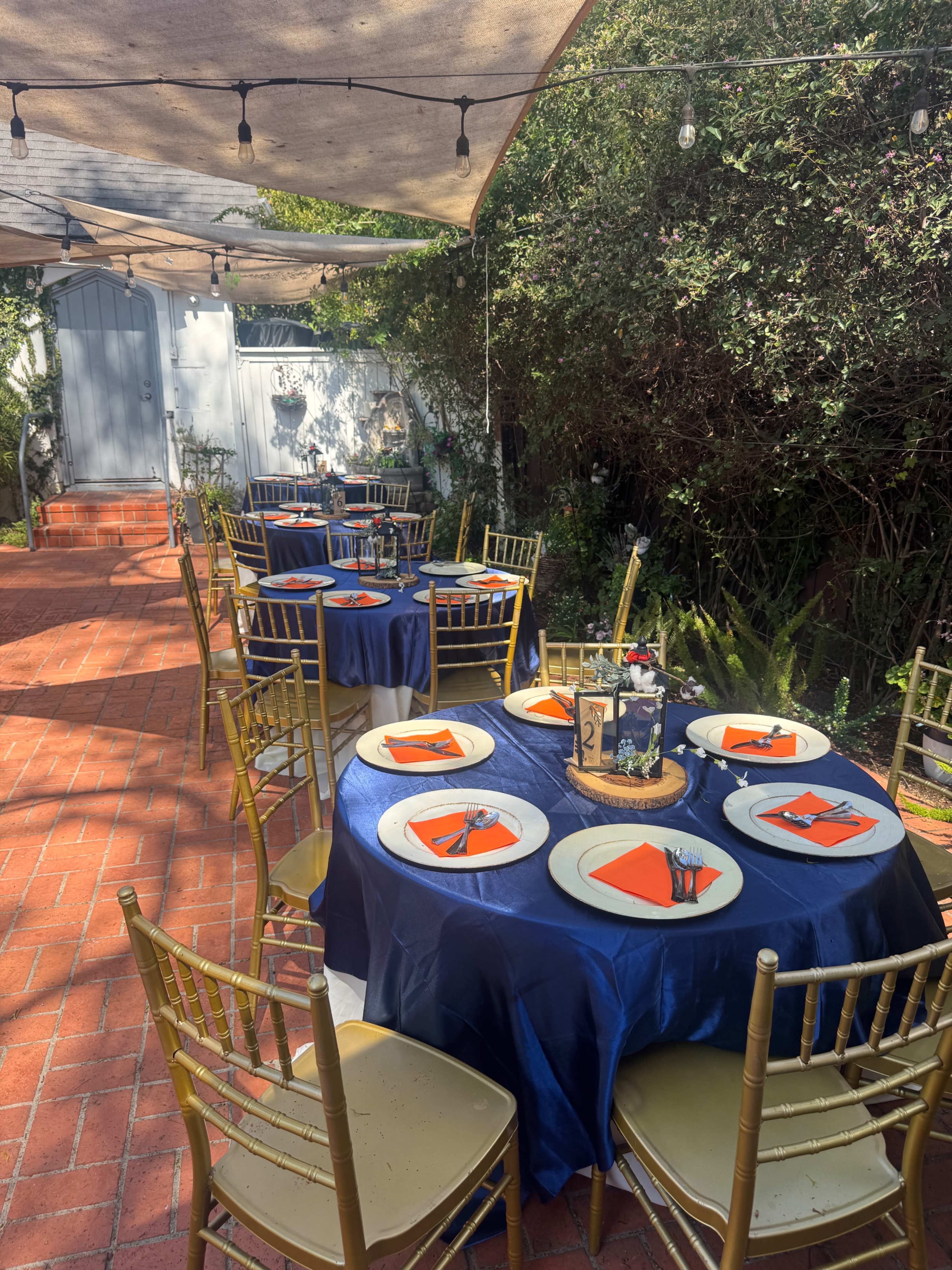 A patio is set up for an outdoor event with round tables covered in blue cloth, each featuring orange napkins and gold chairs.