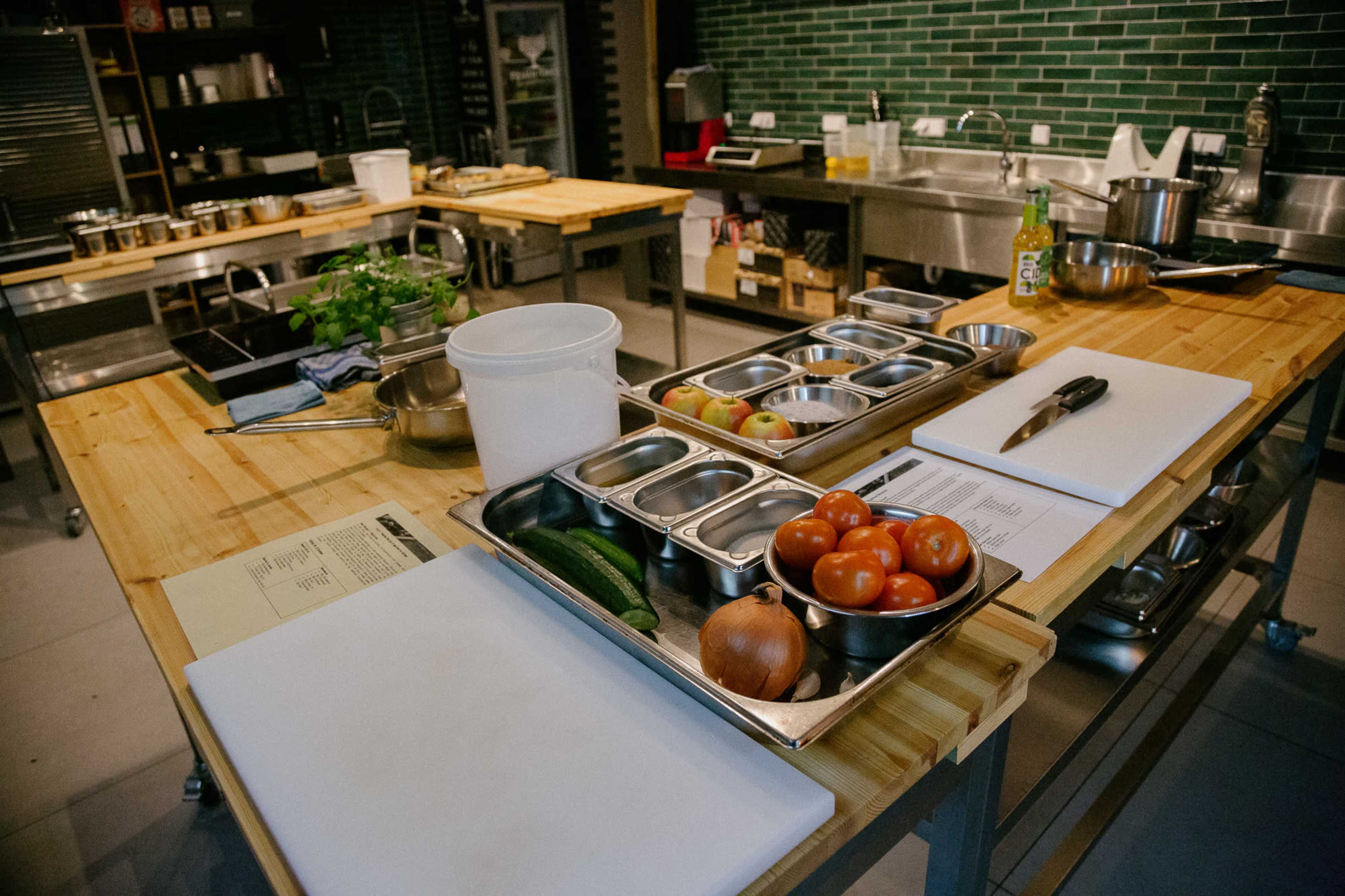 A kitchen workspace features wooden tables with containers of chopped vegetables, utensils, and recipe papers arranged neatly.