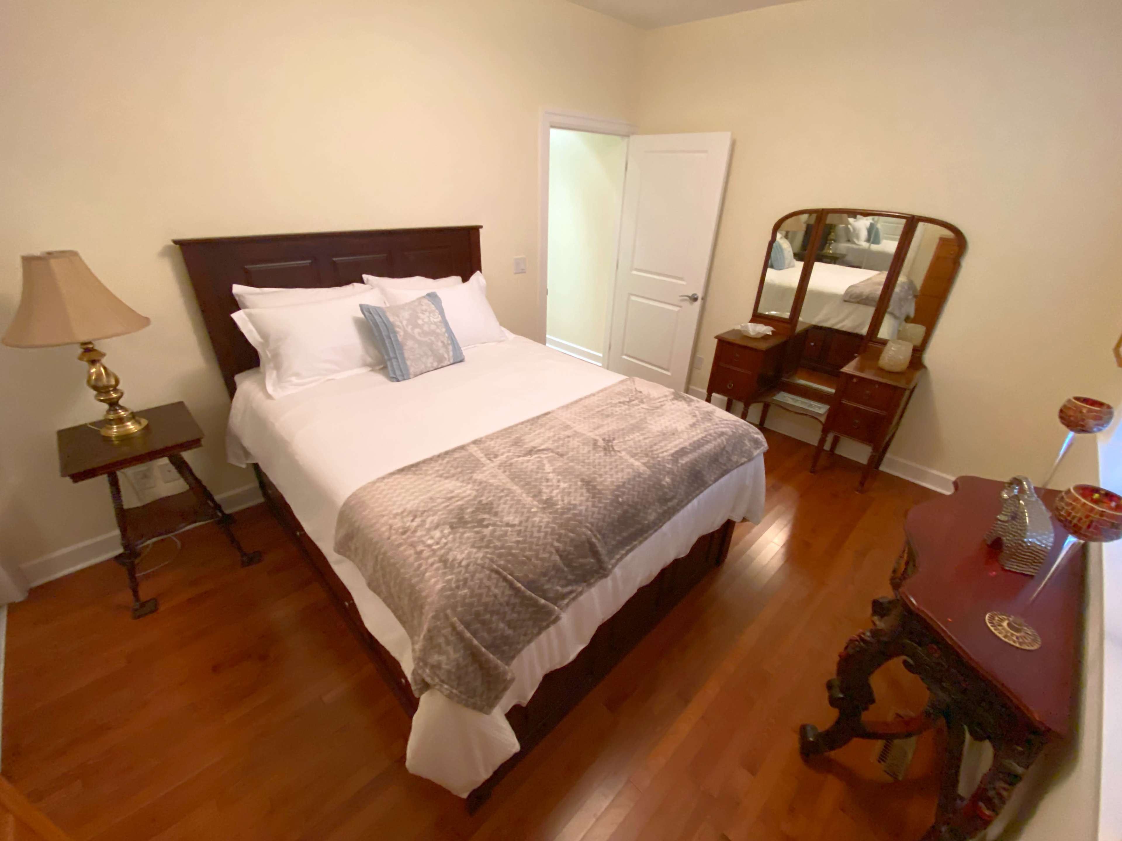 A neatly arranged bedroom featuring a queen-sized bed with white bedding, a wooden nightstand with a lamp, and a dresser with a mirror.