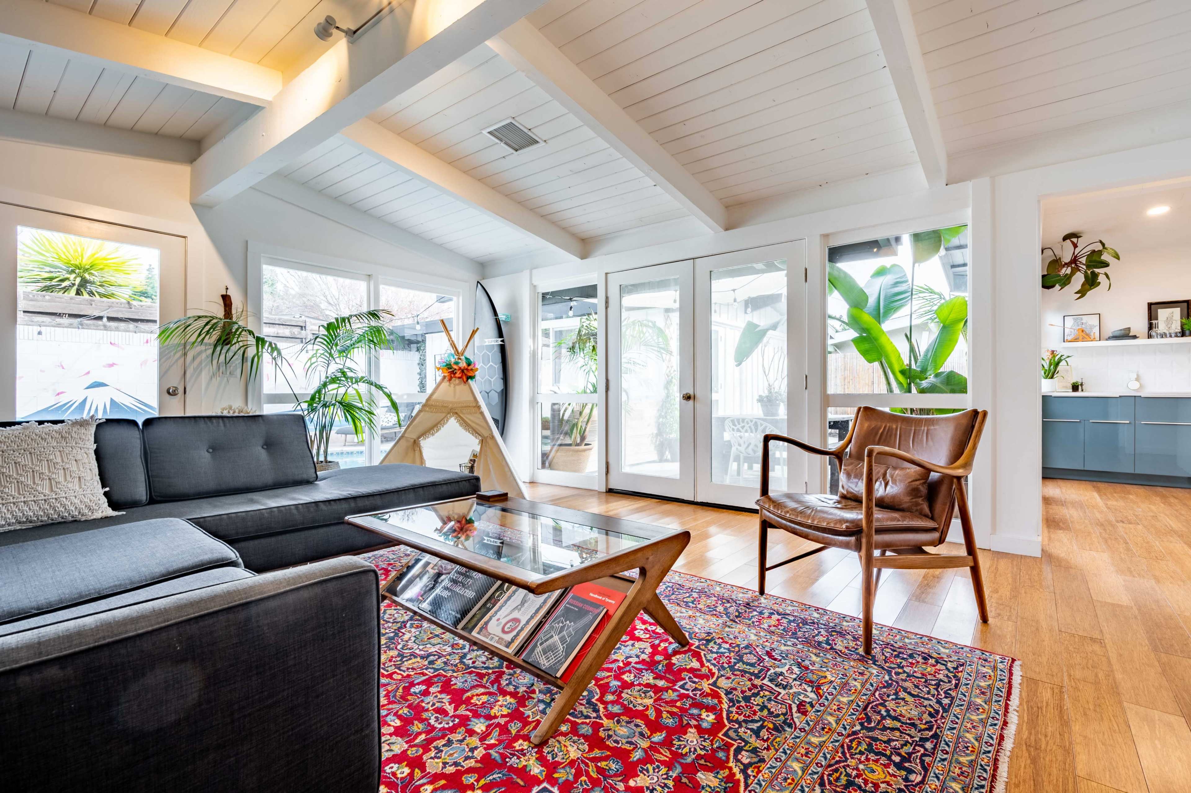 A modern living room with a dark sofa, a glass coffee table on a colorful rug, and large windows that provide natural light and a view of greenery outside.