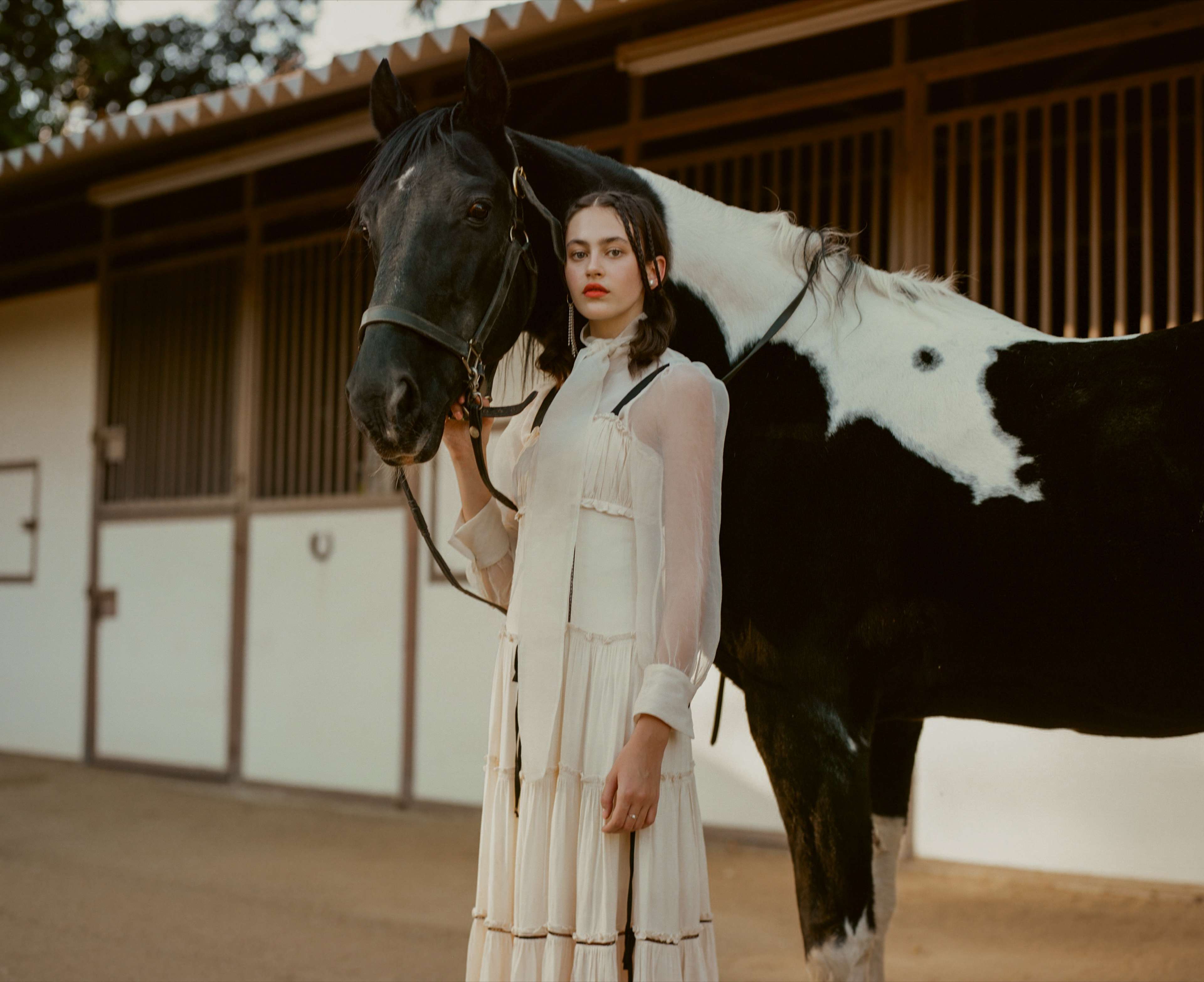 A woman in a white dress stands next to a black and white horse in a barn setting.