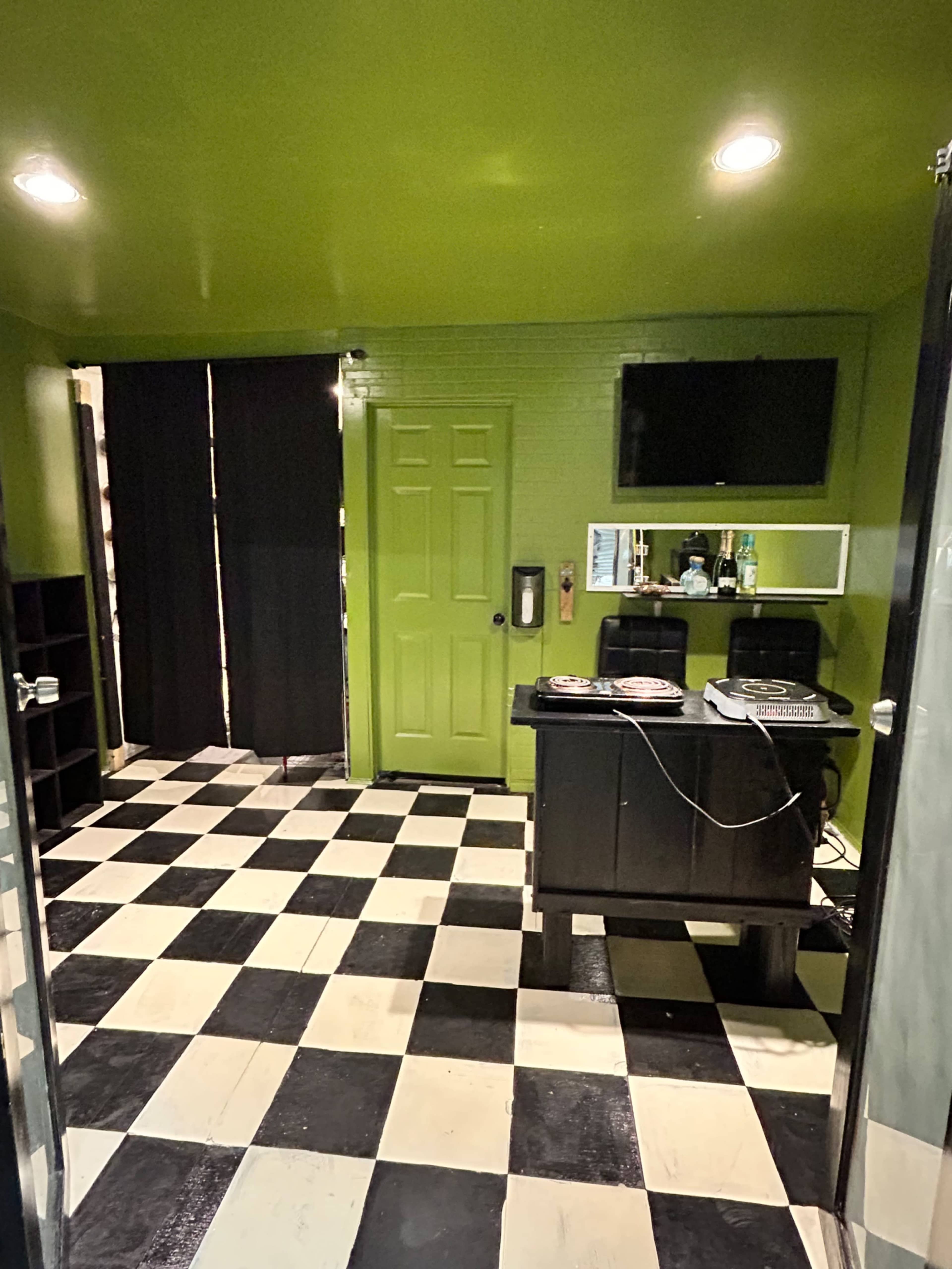 The image shows a room with a green ceiling and walls, featuring a black and white checkered floor, a small table with equipment, a TV on the wall, and a black curtain covering a doorway.