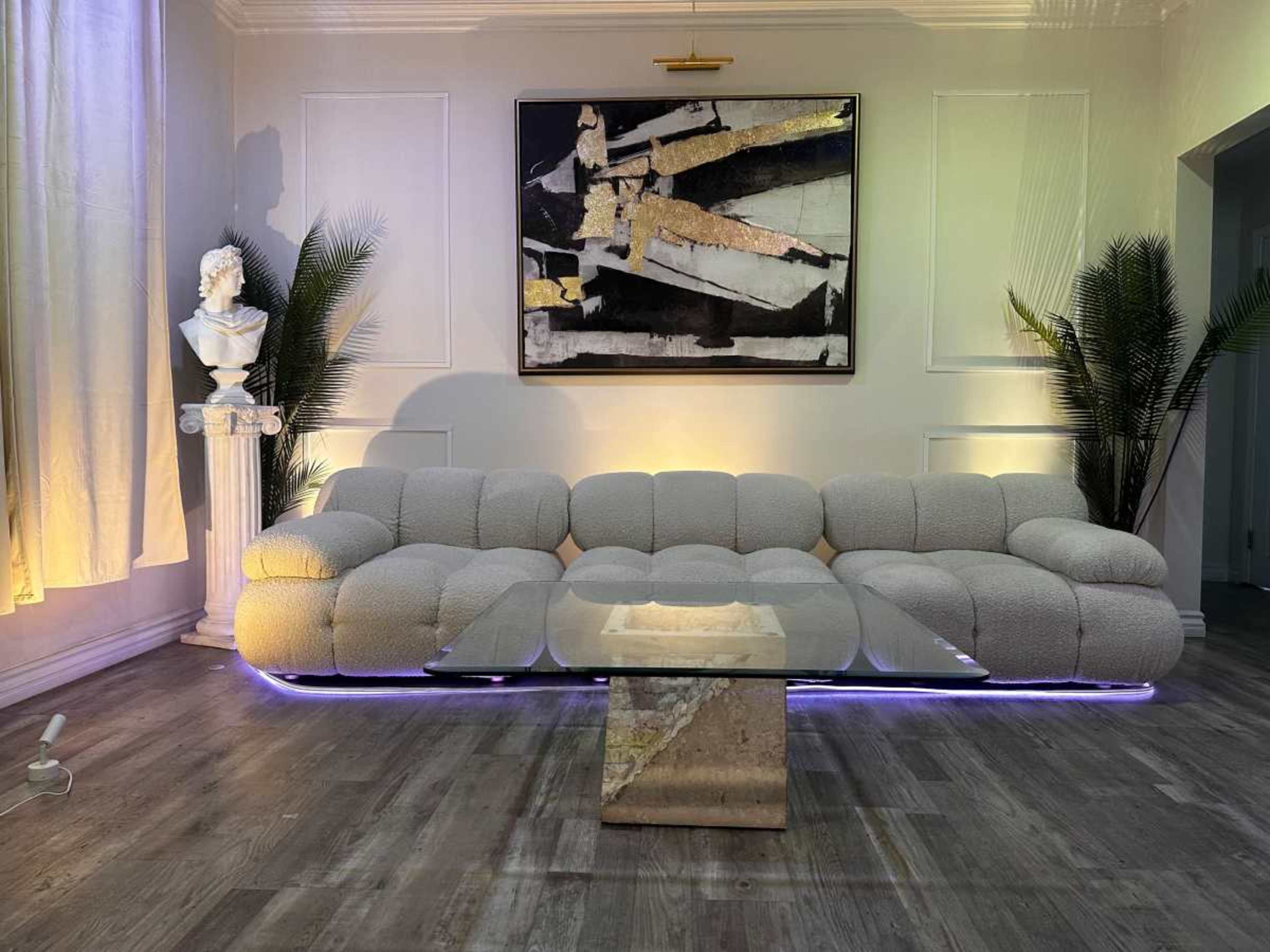 The image shows a modern living room featuring a plush, cream-colored sectional sofa, a glass coffee table with a marble base, a large abstract painting on the wall, and decorative plants.