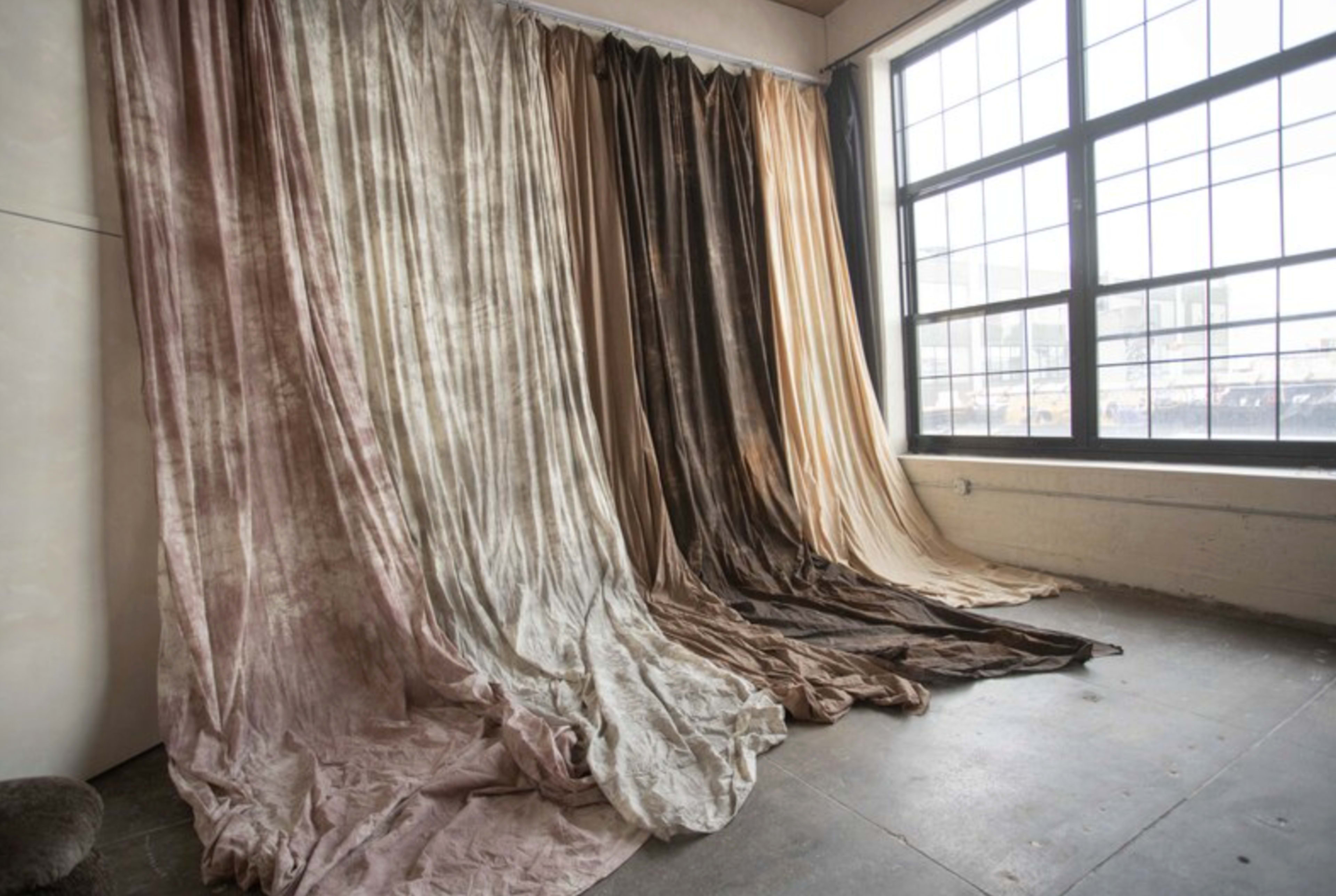 The image shows a large window with multiple draped fabrics in varying shades, cascading down the wall in a studio-like space.