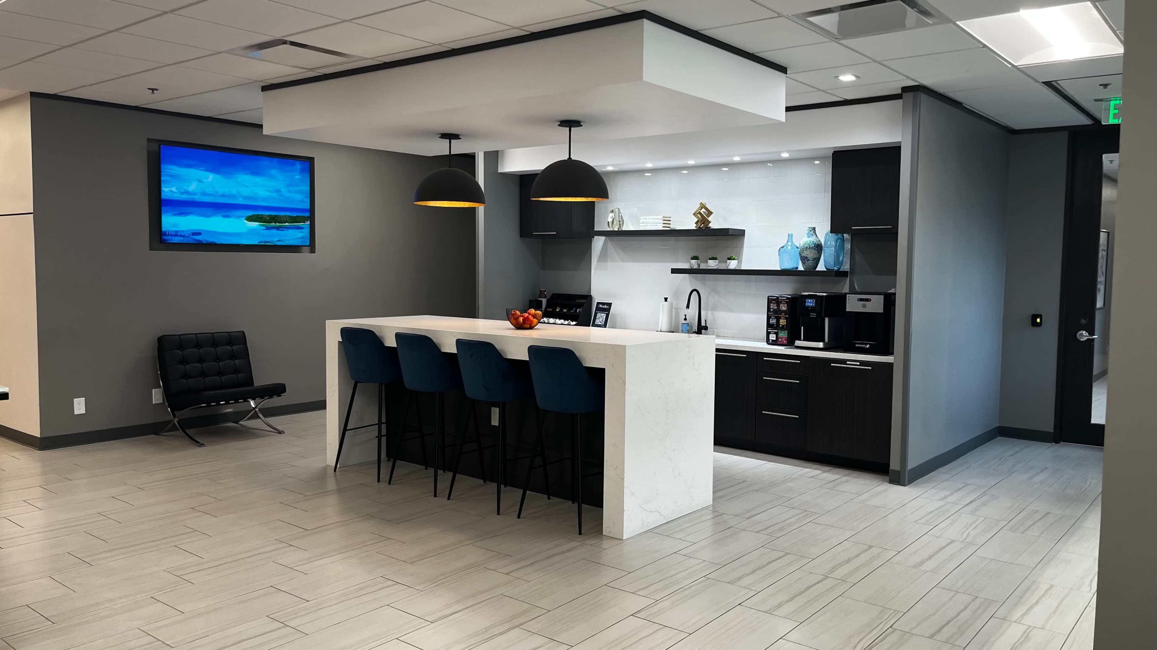 The image shows a modern office kitchenette featuring a white island counter with four blue bar stools, black pendant lights overhead, and a wall-mounted TV displaying a nature scene.