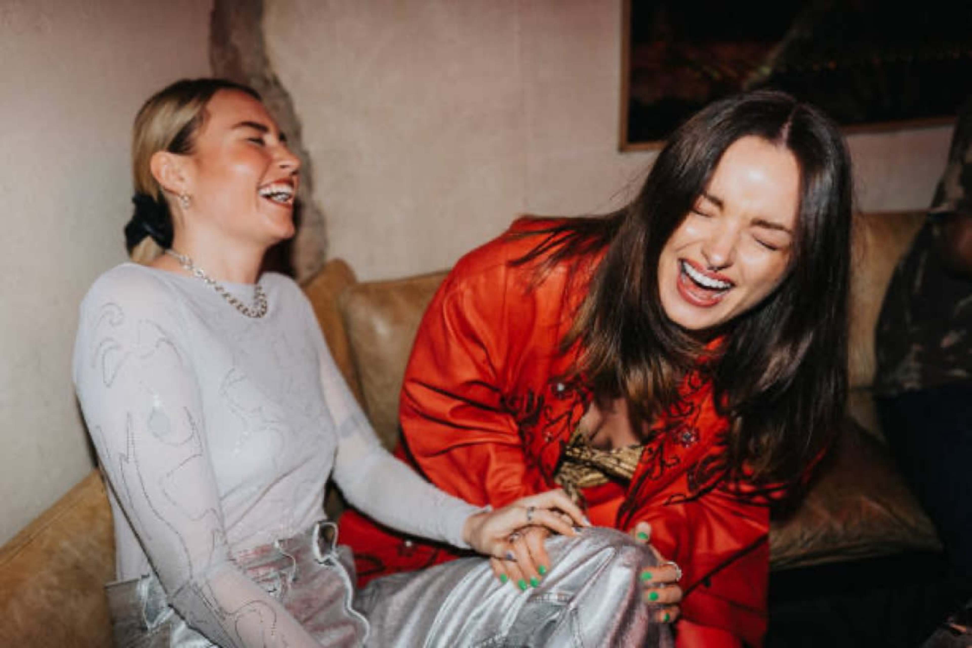 Two women are seated on a couch laughing together in a social setting, with one wearing a silver outfit and the other in an orange coat.
