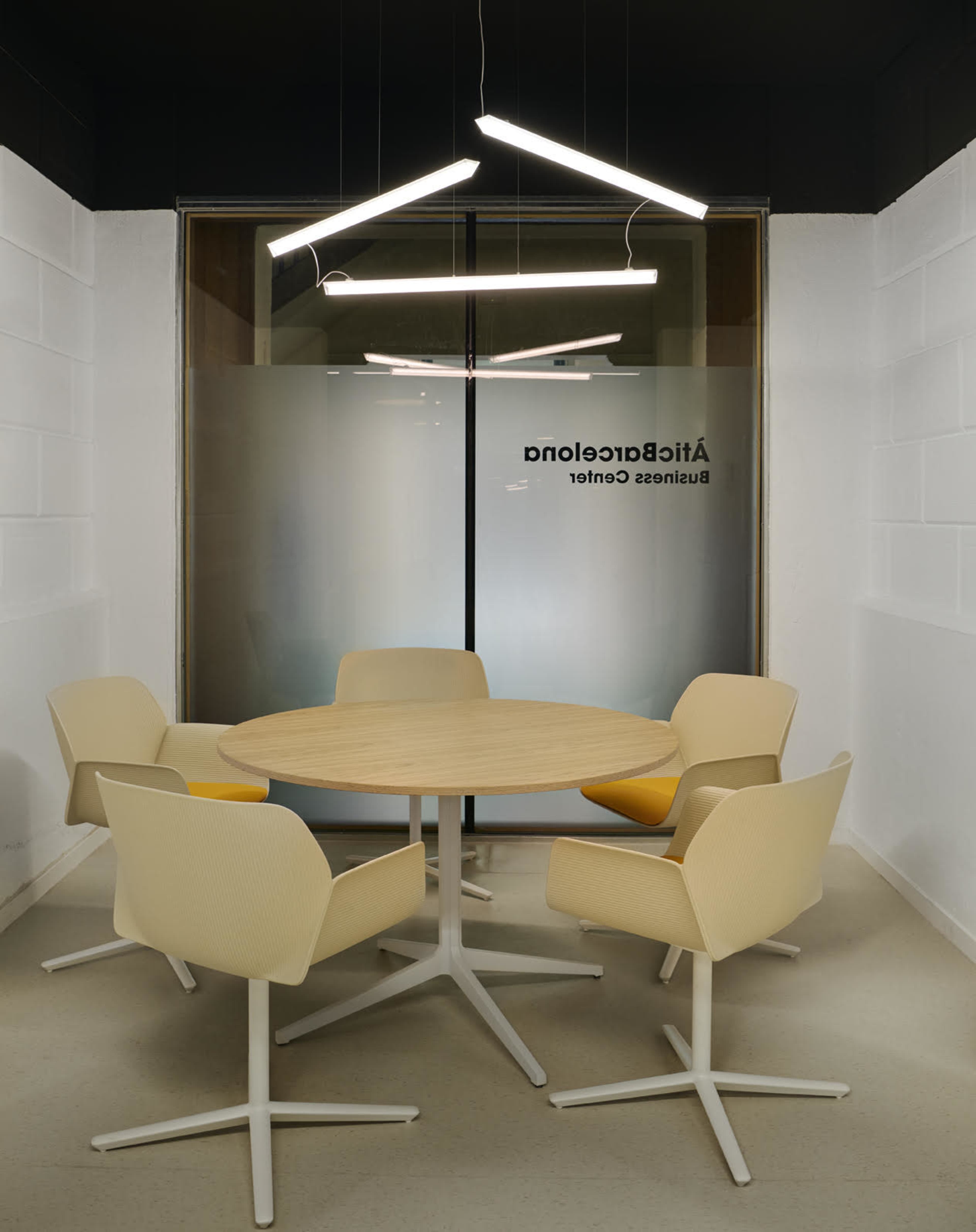 A round wooden table surrounded by four beige chairs is set in a modern conference room with a glass wall.