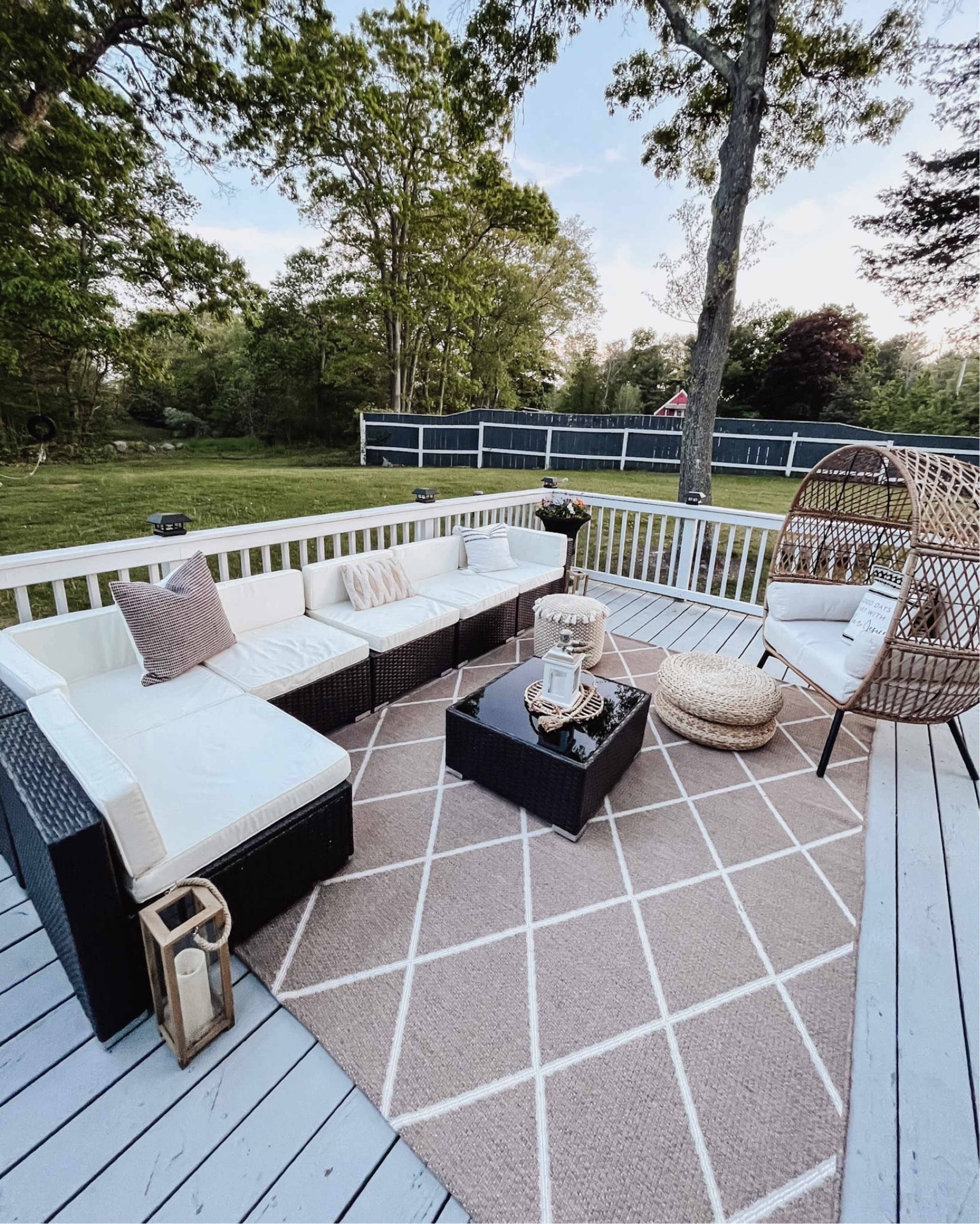 A patio area featuring a sectional couch, a round chair, a coffee table on a geometric-patterned rug, and surrounded by trees and a grassy yard.