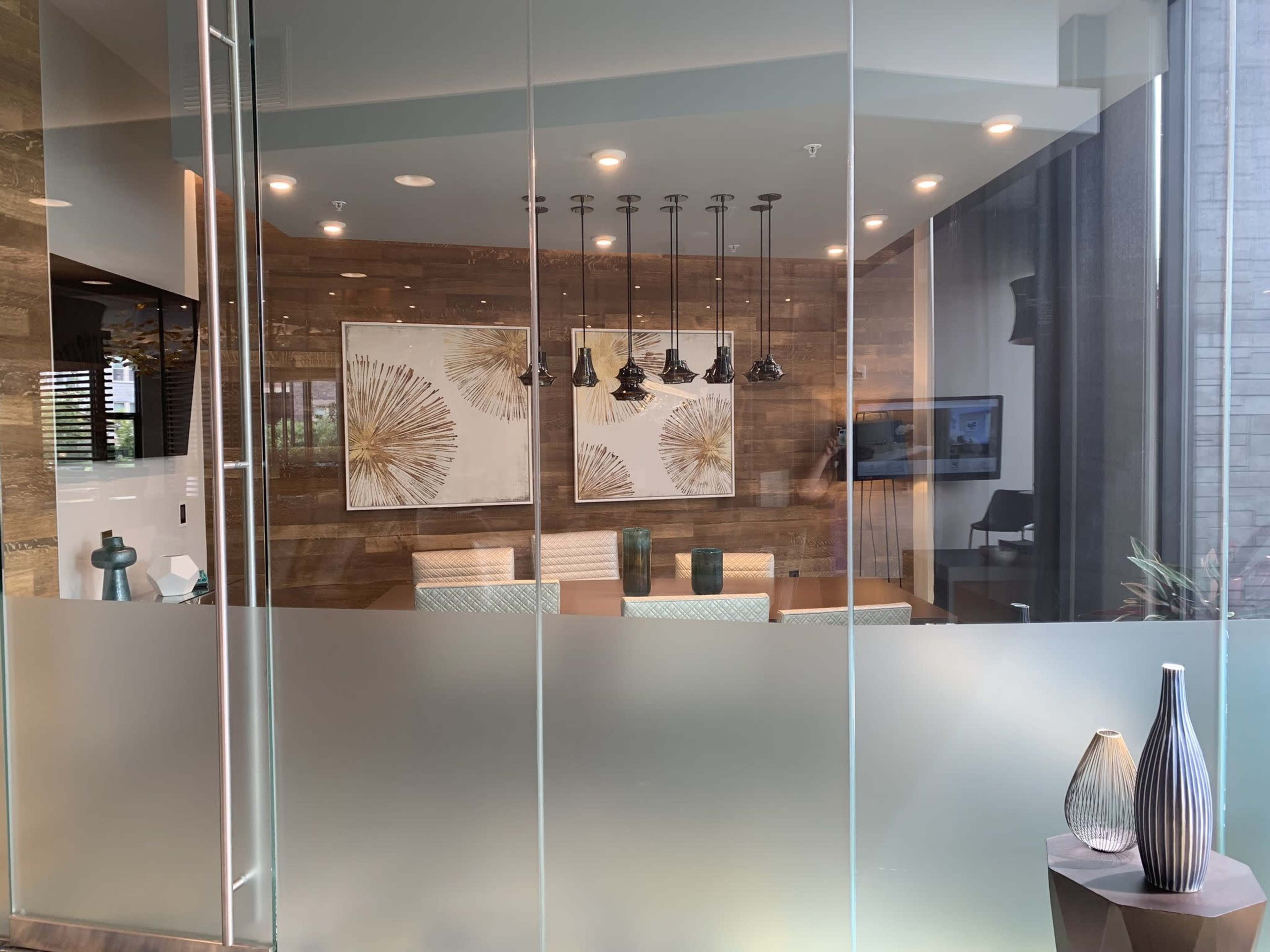 A modern office meeting room with large glass walls, featuring two framed artworks, pendant lights, and a seating area with upholstered chairs.