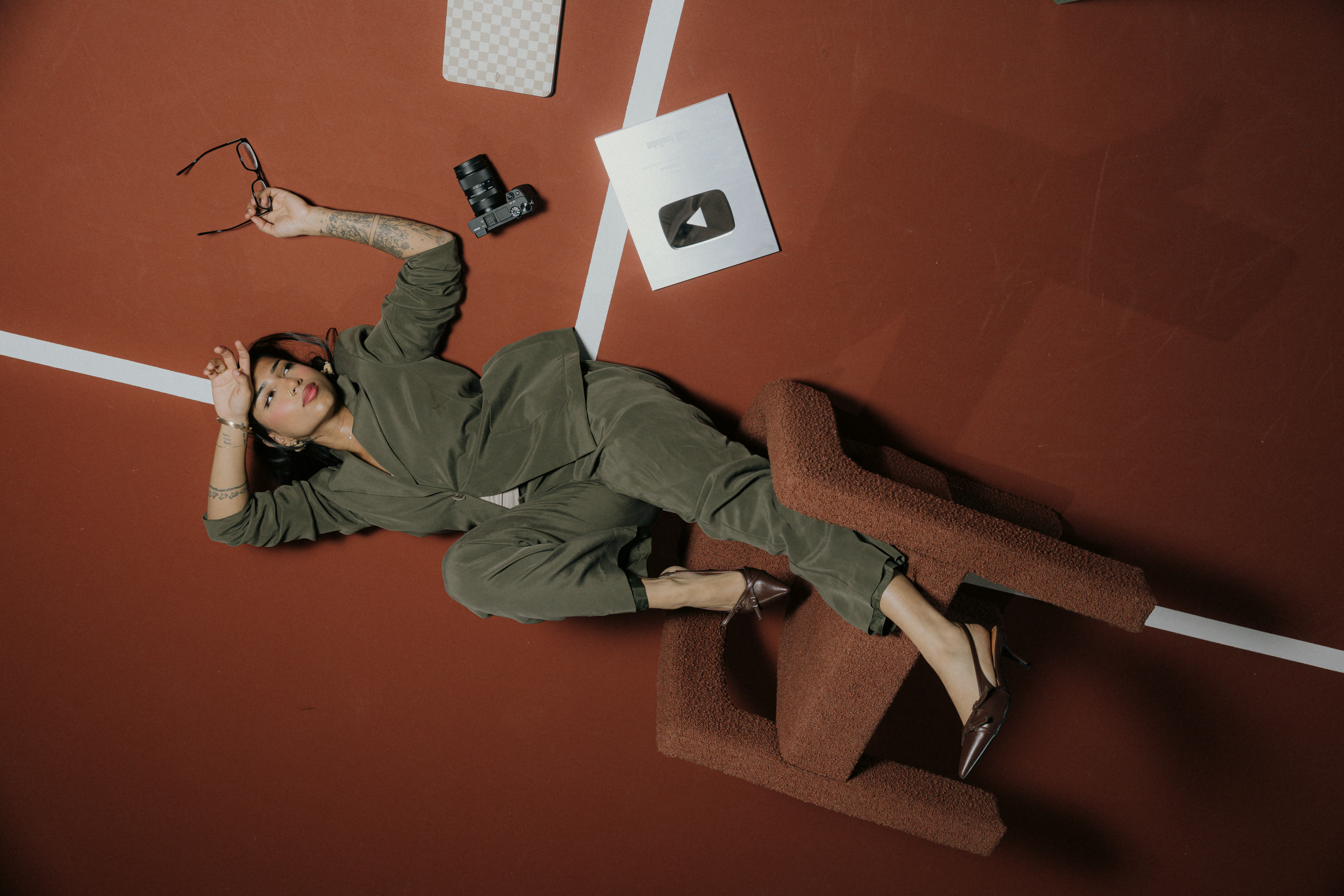 A woman in a green outfit lies on a red floor surrounded by a camera, glasses, and a YouTube play button plaque.