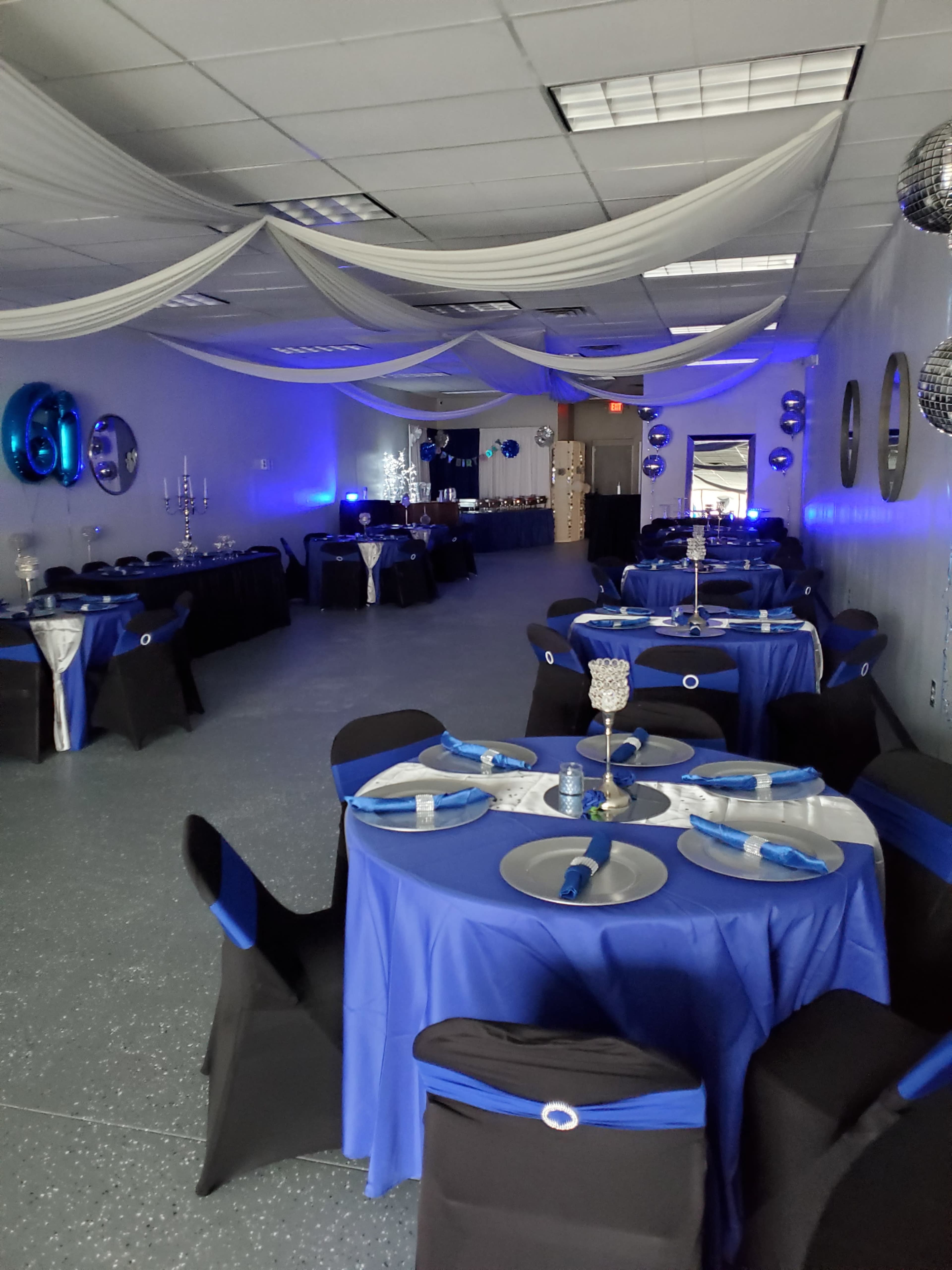 A decorated event space with tables covered in blue tablecloths and black chairs, set up for a celebration with blue and silver accents.