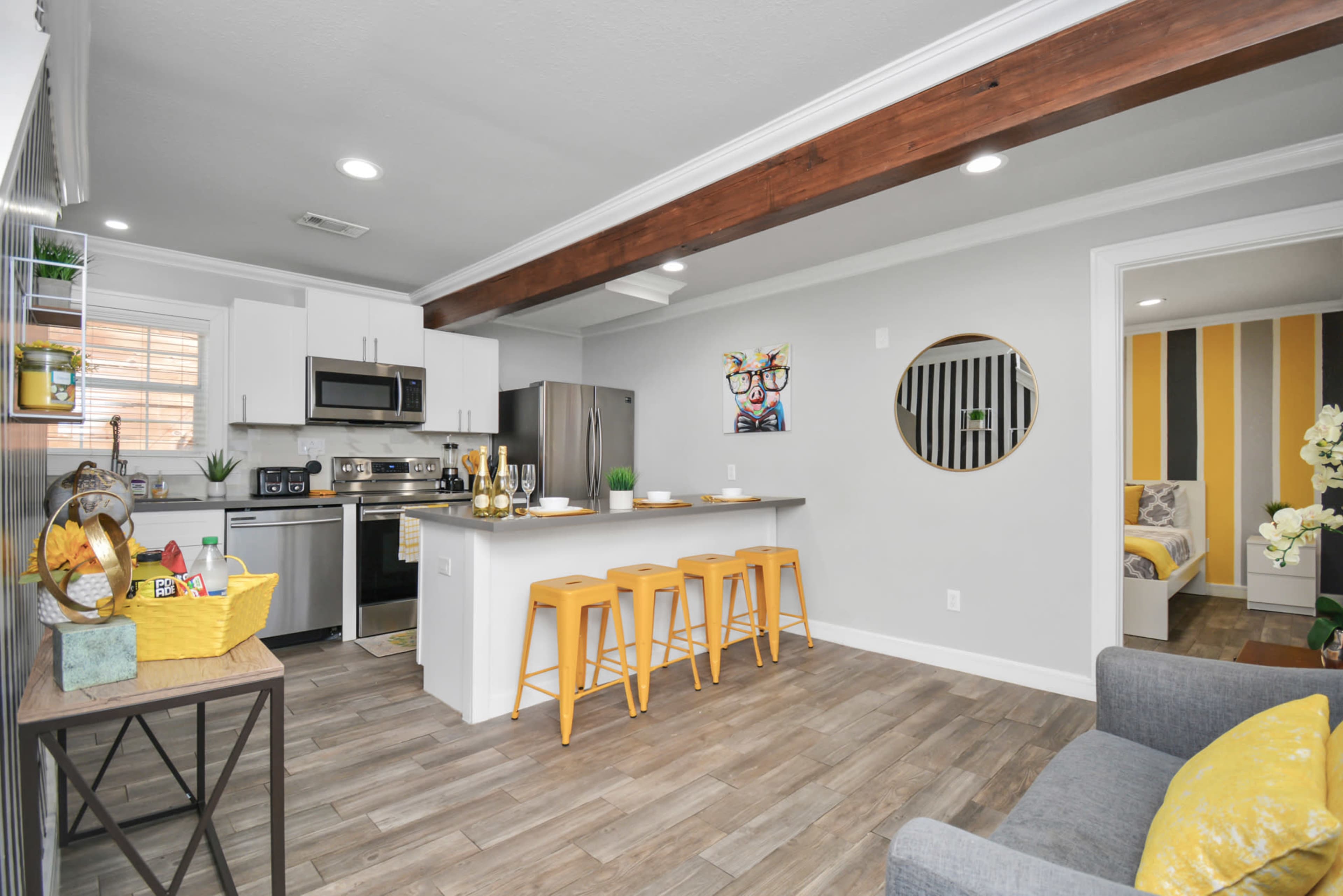 A modern kitchen and living area feature stainless steel appliances, a wooden beam ceiling, yellow bar stools, and a view into a bedroom with yellow and gray decor.