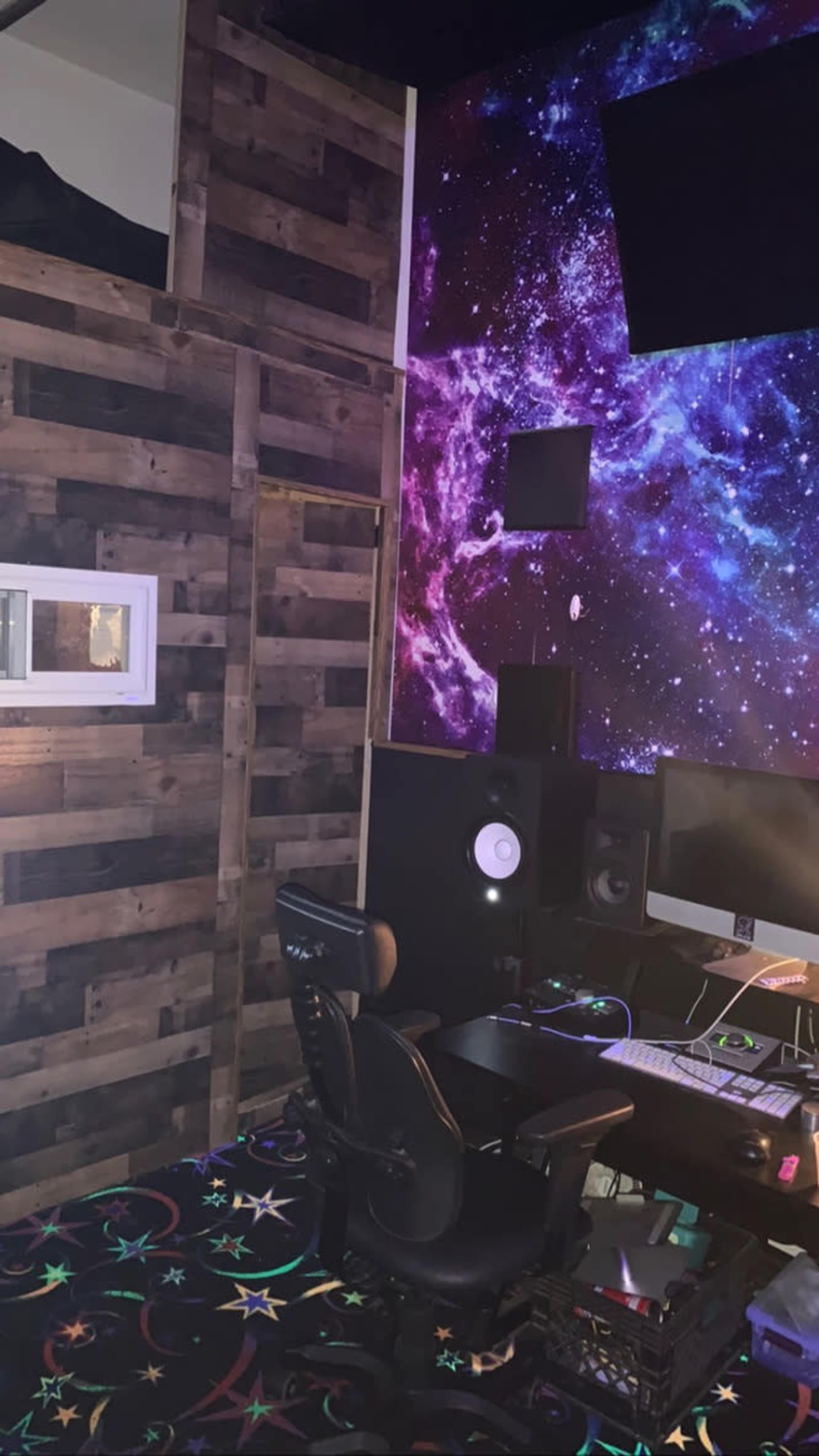 A room with a wooden wall, a cosmic-themed mural, a computer workstation with audio equipment, and a colorful floor.