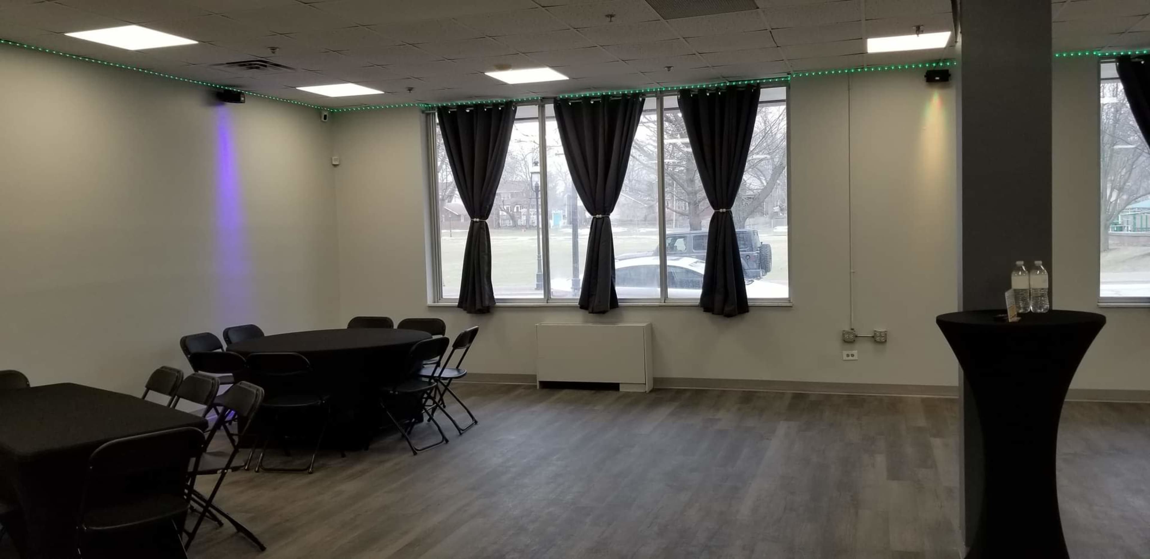 The room features several black round tables with chairs, large windows draped in dark curtains, and a tall black table with bottles of water against a light-colored wall.