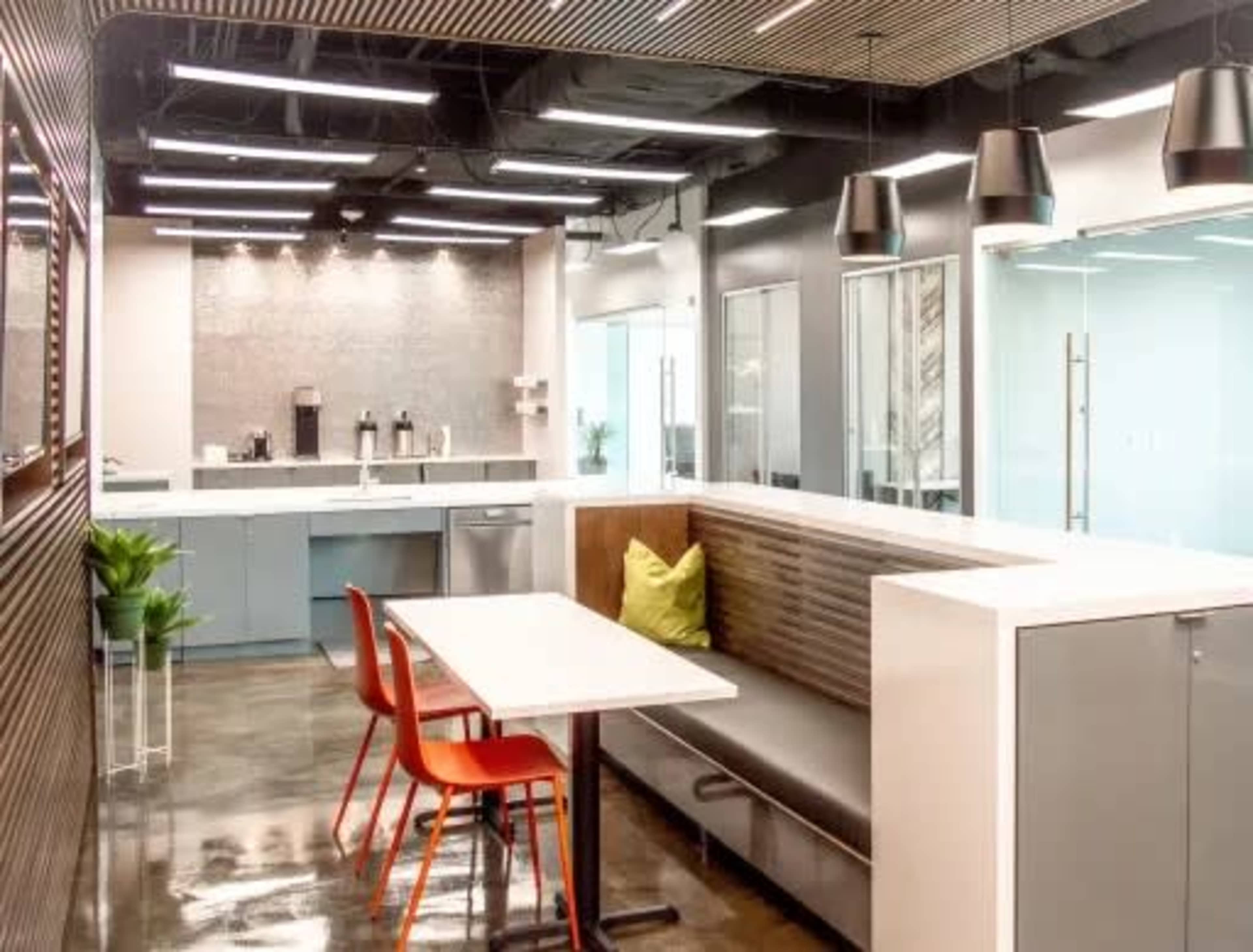 The image shows a modern office break room with a kitchenette, seating area, and a polished concrete floor.