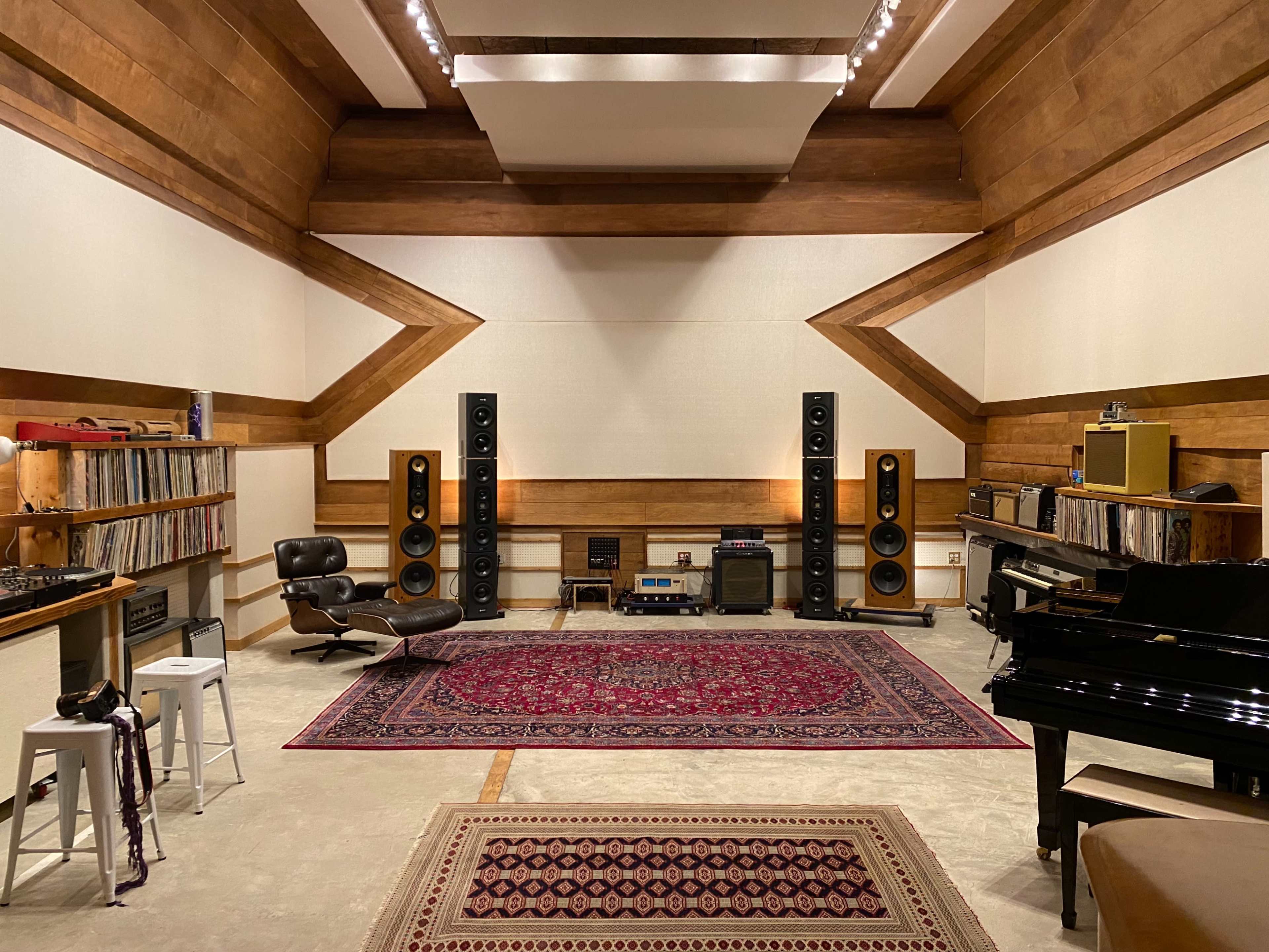 The image shows a spacious audio studio with wooden walls, large speakers, a grand piano, and a collection of vinyl records displayed on shelves.
