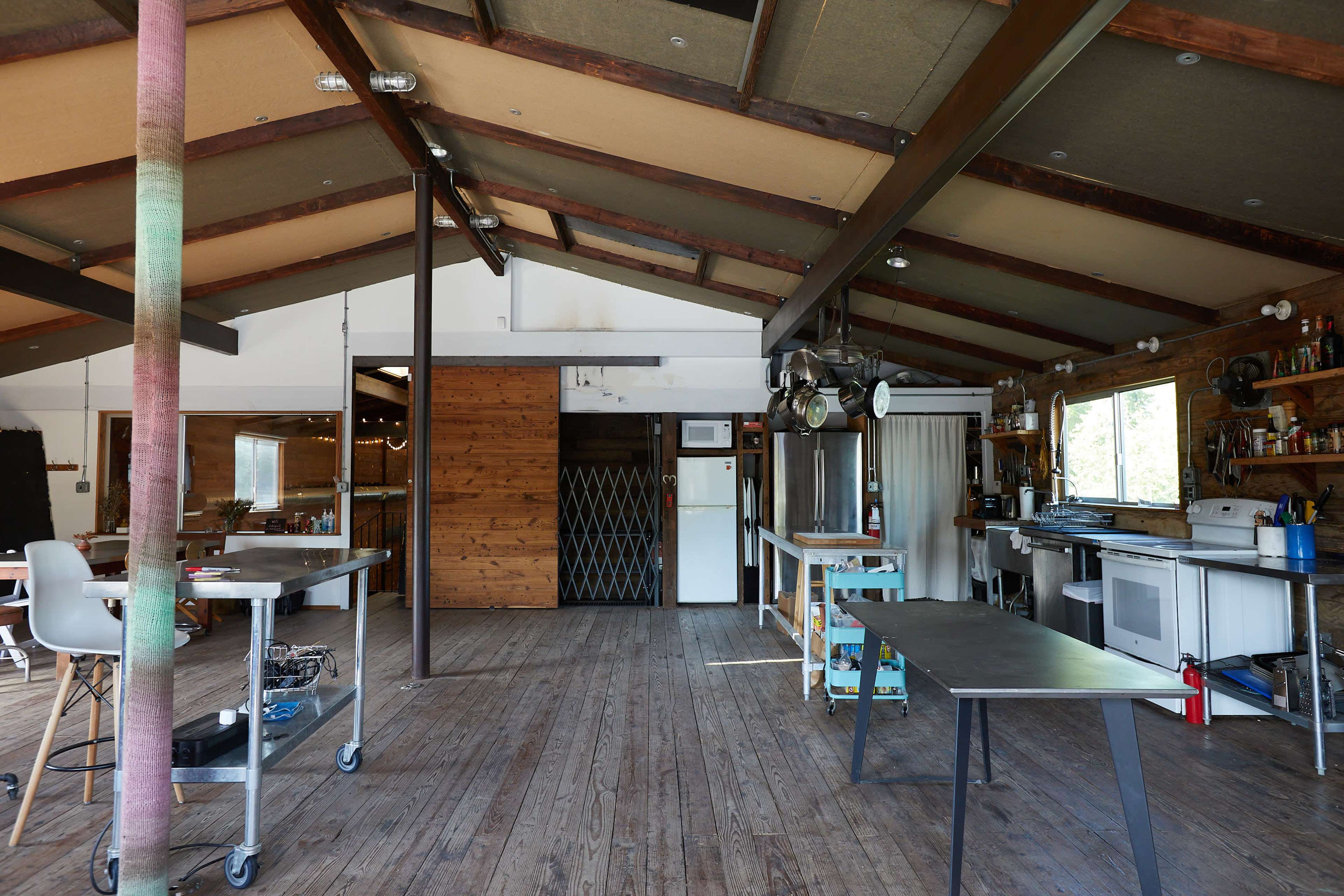 An open interior space with wooden flooring, exposed beams, and various kitchen appliances and workspaces arranged throughout.