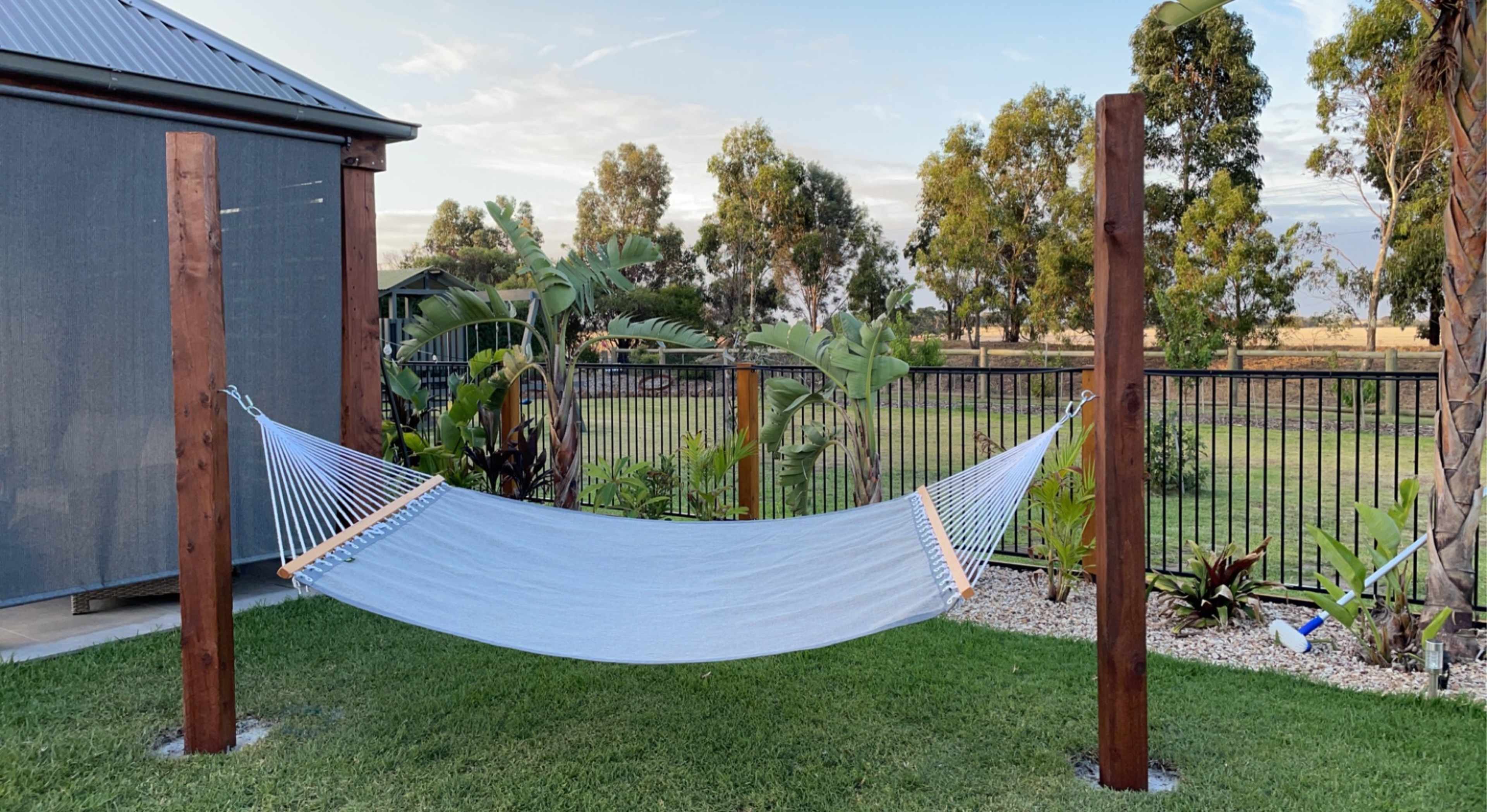 A hammock is suspended between two wooden posts in a grassy backyard surrounded by trees and plants.
