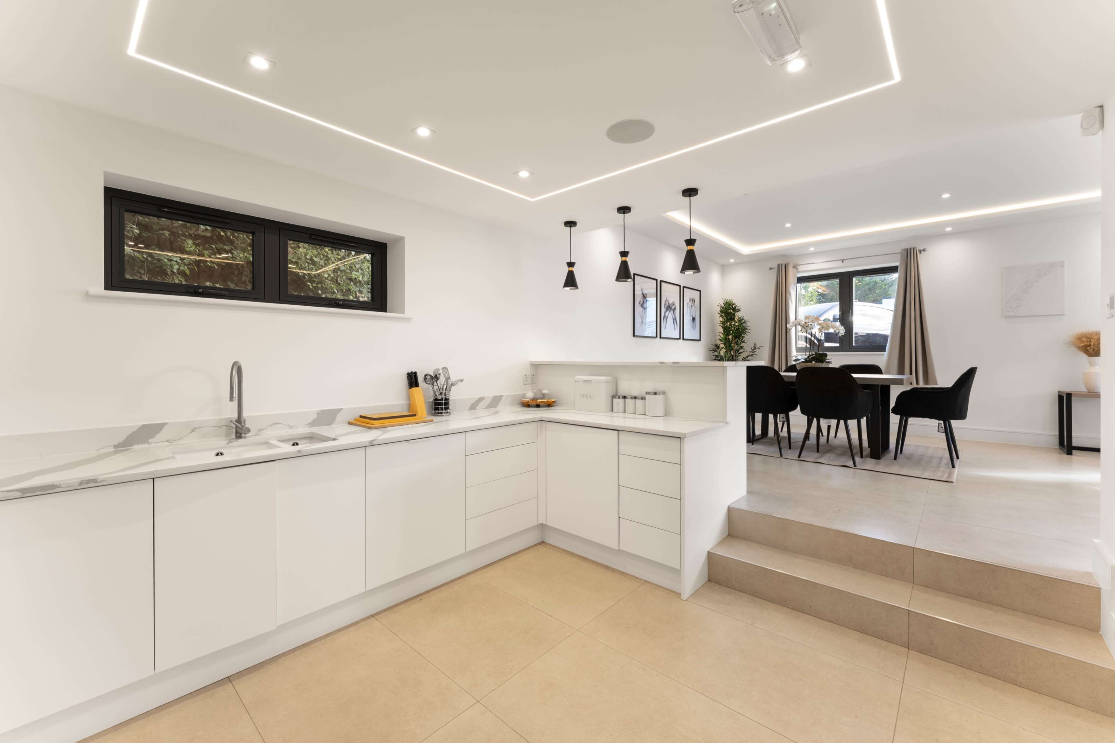 The image shows a modern kitchen with white cabinetry, a marble countertop, and an open dining area with a small table and chairs nearby.