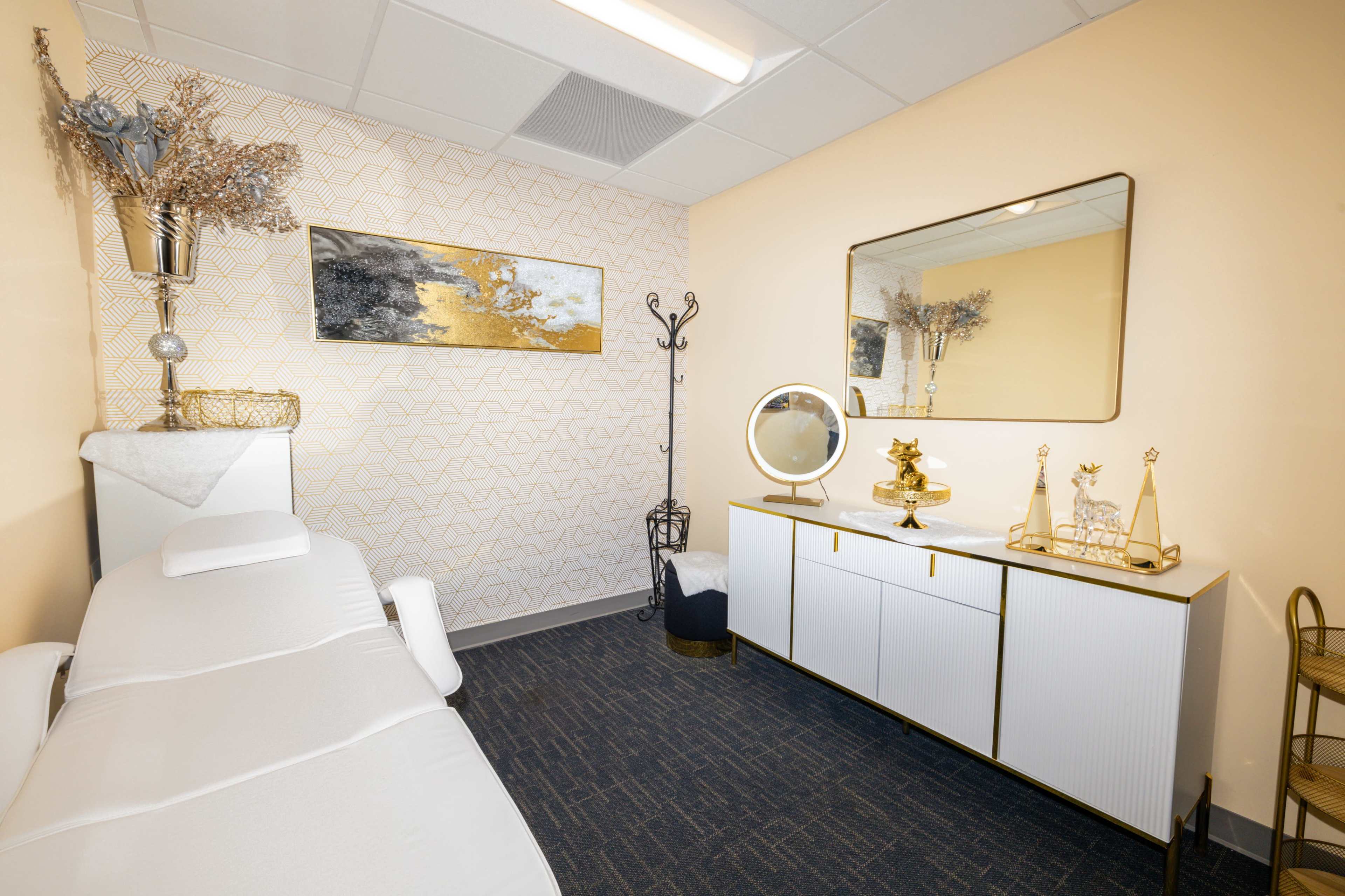 The image shows a minimalist treatment room featuring a massage table, a decorative mirror, a small cabinet, and a wall adorned with geometric art.