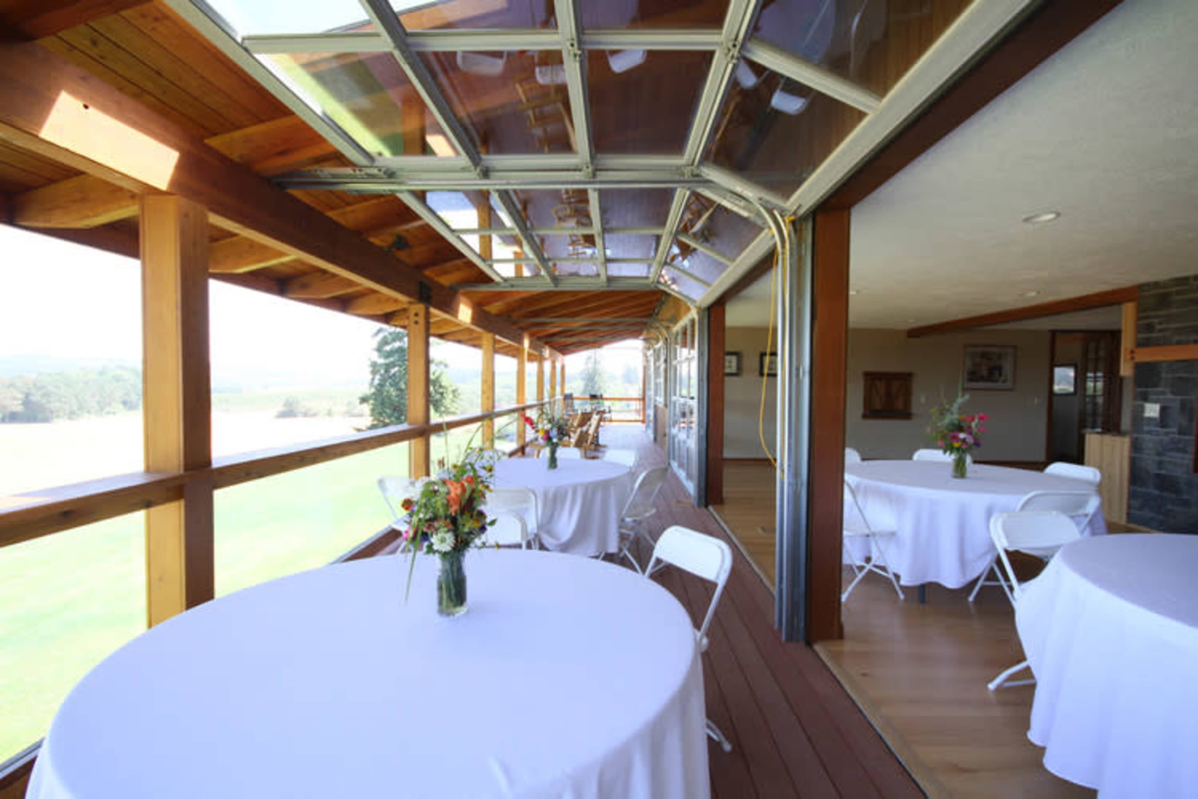 An interior space with several round tables covered in white tablecloths, surrounded by large windows that overlook a grassy area.