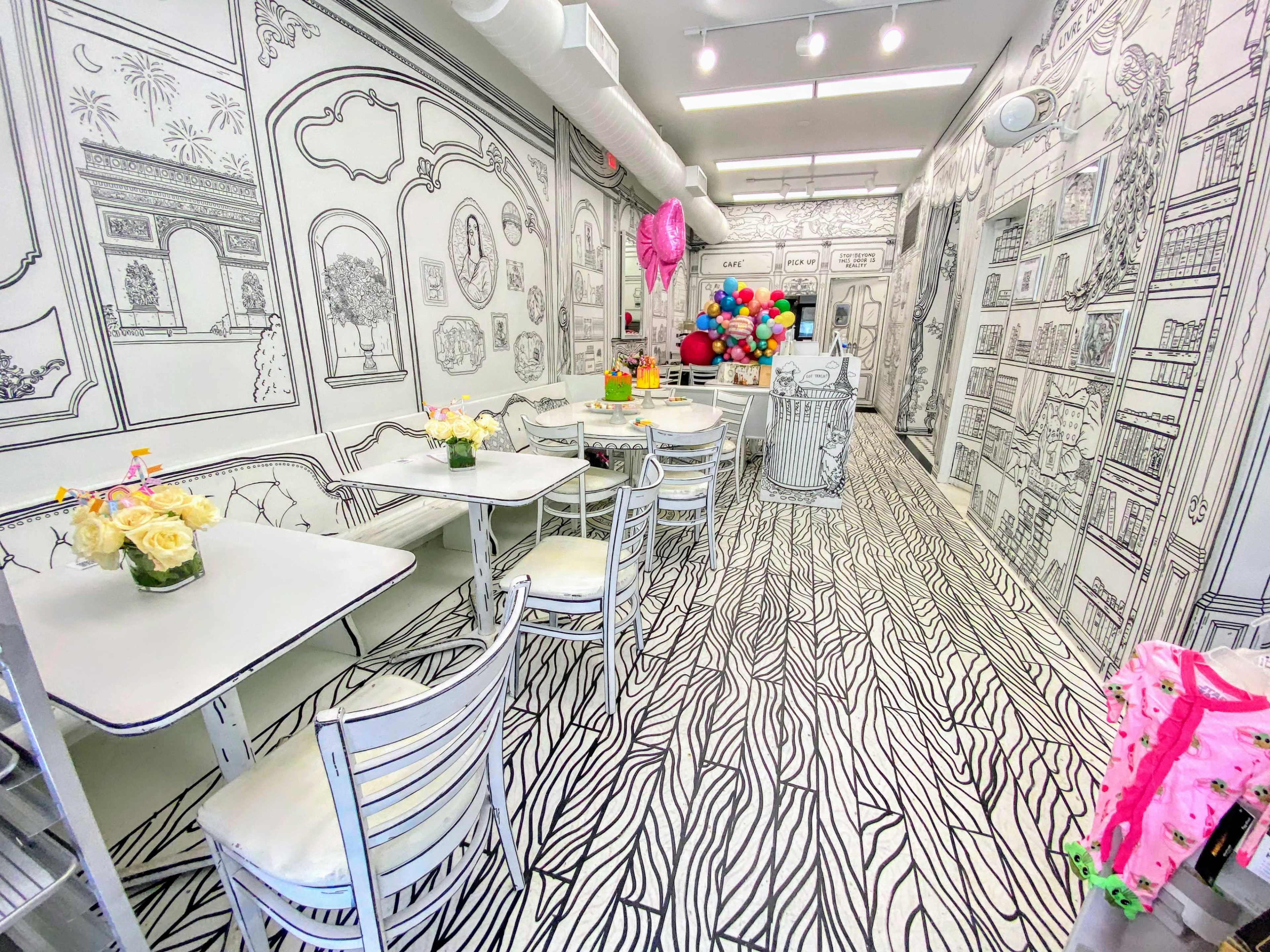 A brightly lit café with black and white illustrated walls, floor, and furniture, featuring tables and chairs arranged neatly with floral decorations.