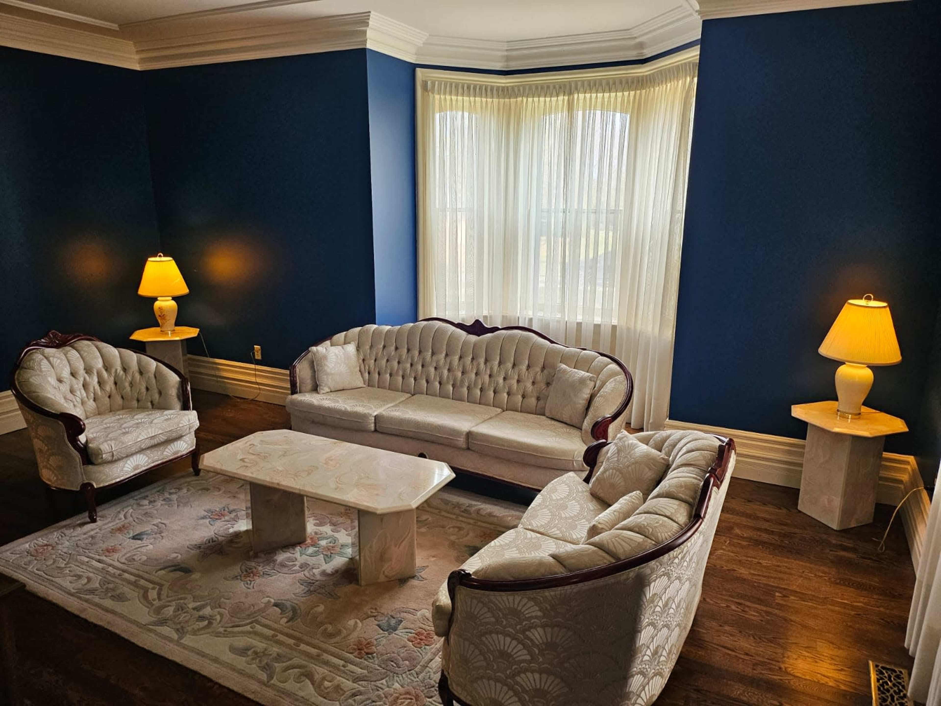 A beautifully furnished living room with a light-colored upholstery sofa set, a marble coffee table, and two lamps illuminating the space against a dark blue wall.