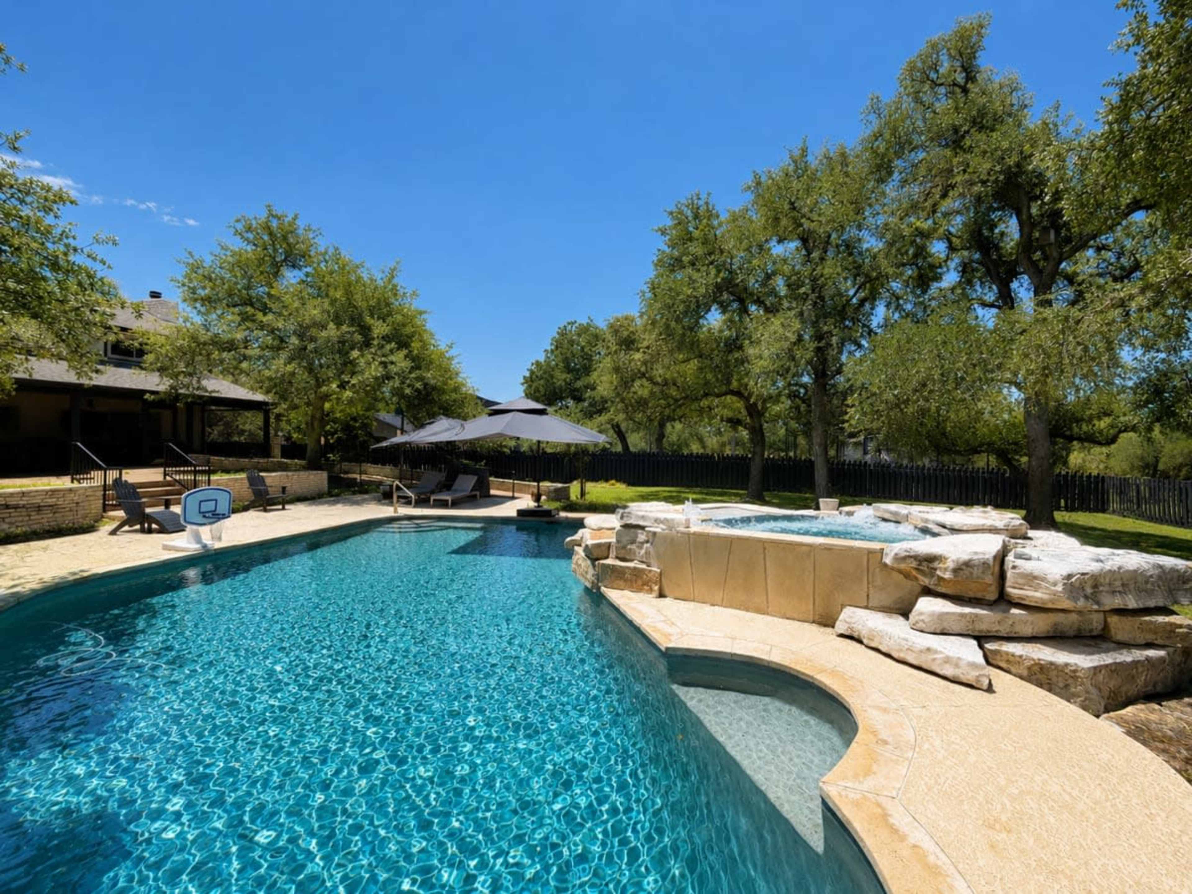The image shows a clear blue swimming pool surrounded by sandy areas and lounge chairs, with trees and a patio in the background.