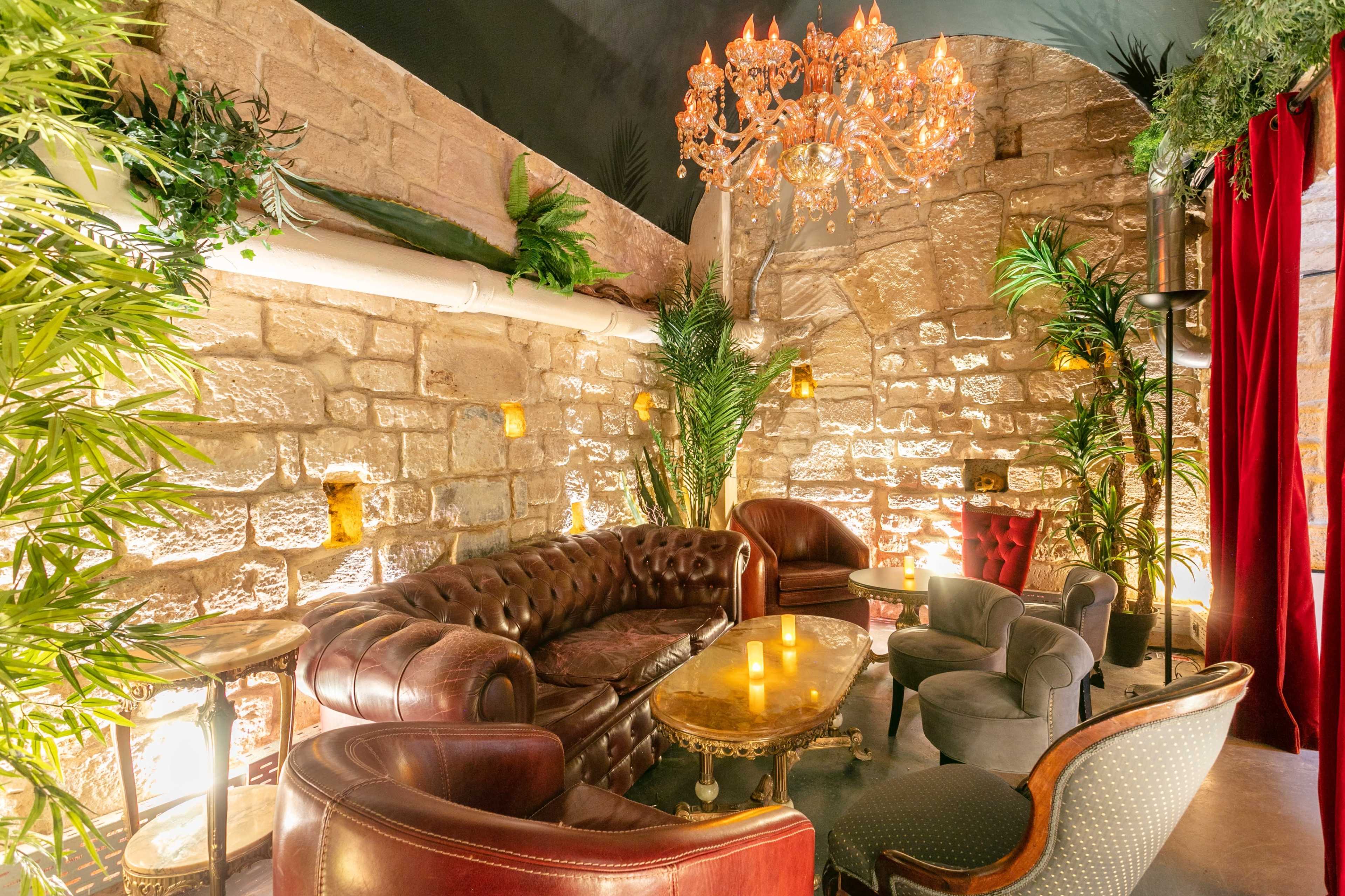 A cozy lounge area with a plush leather sofa, elegant chairs, and decorative plants against a backdrop of stone walls and a chandelier.