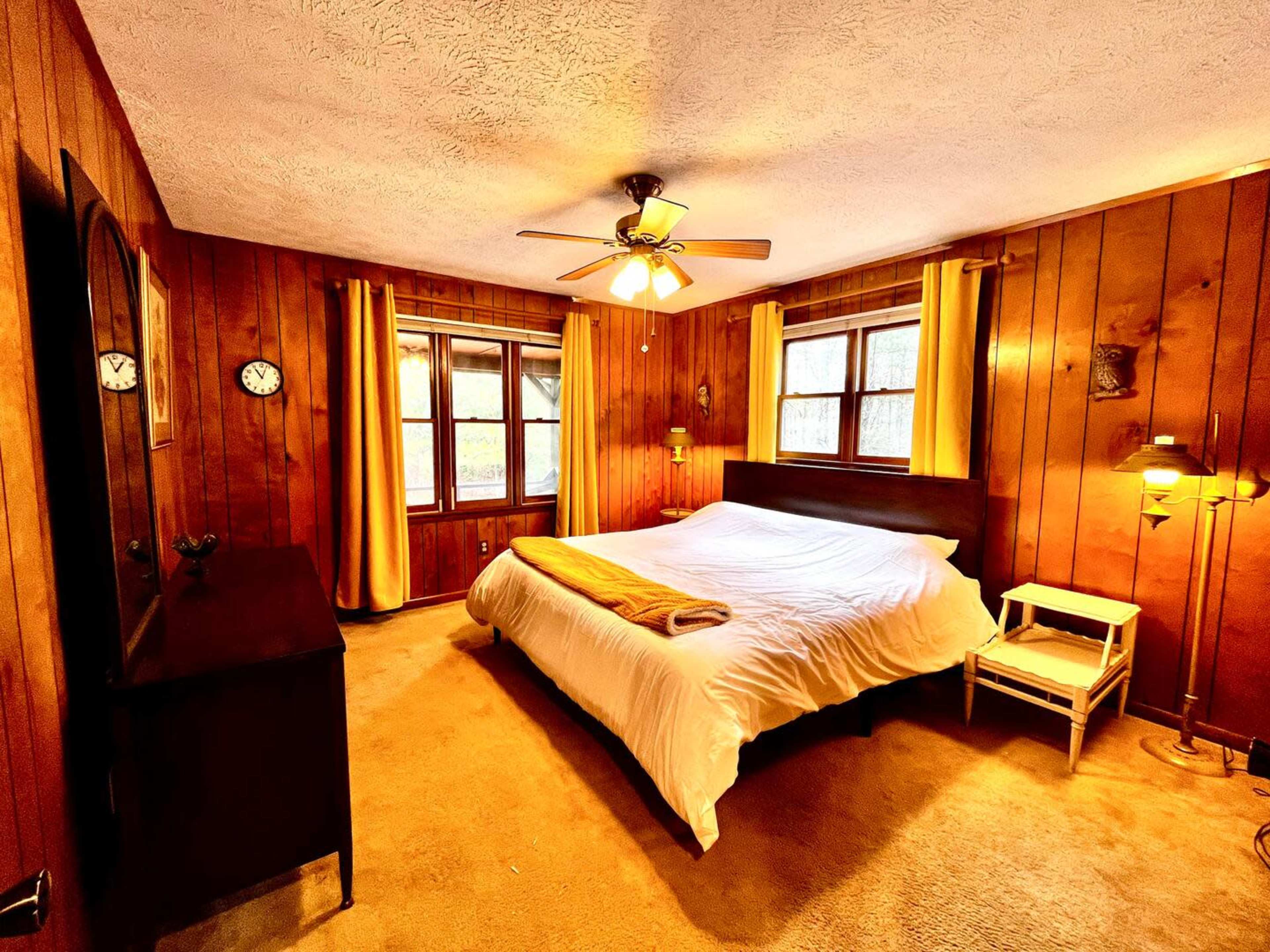 A wooden-paneled bedroom featuring a large bed, two windows with yellow curtains, and a dresser beside a small side table with a lamp.