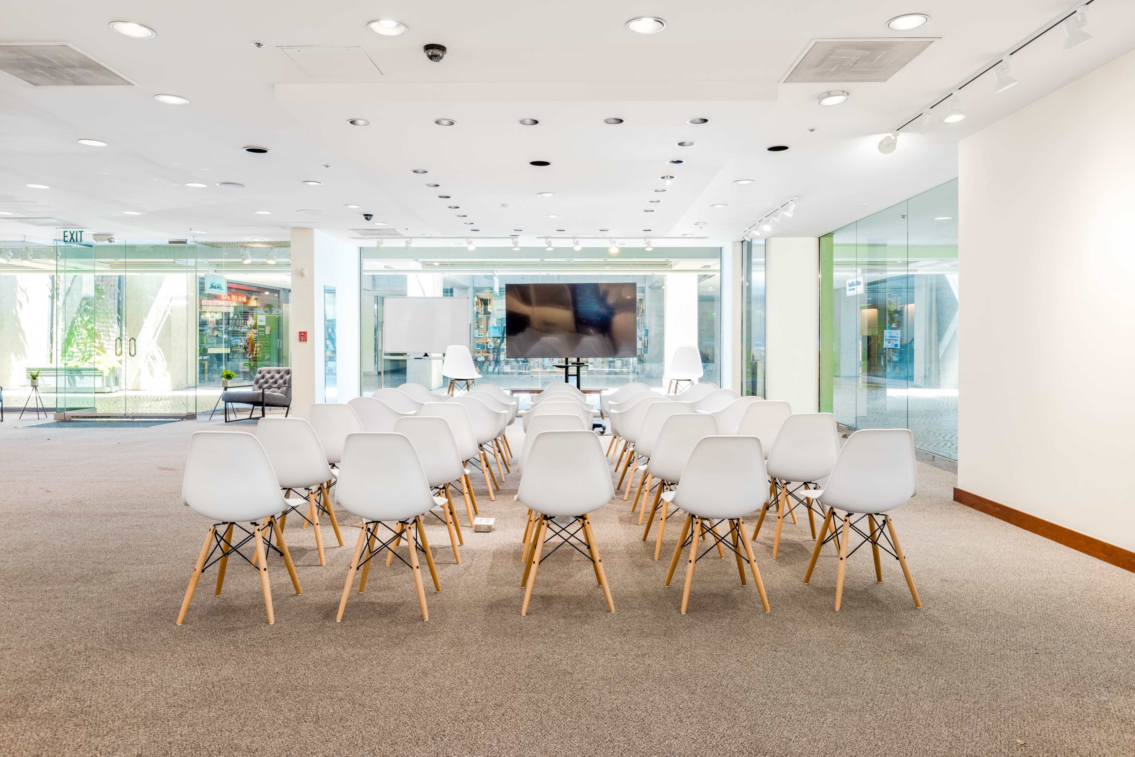 A row of white chairs with wooden legs is set up facing a large screen in a brightly lit room with glass walls.