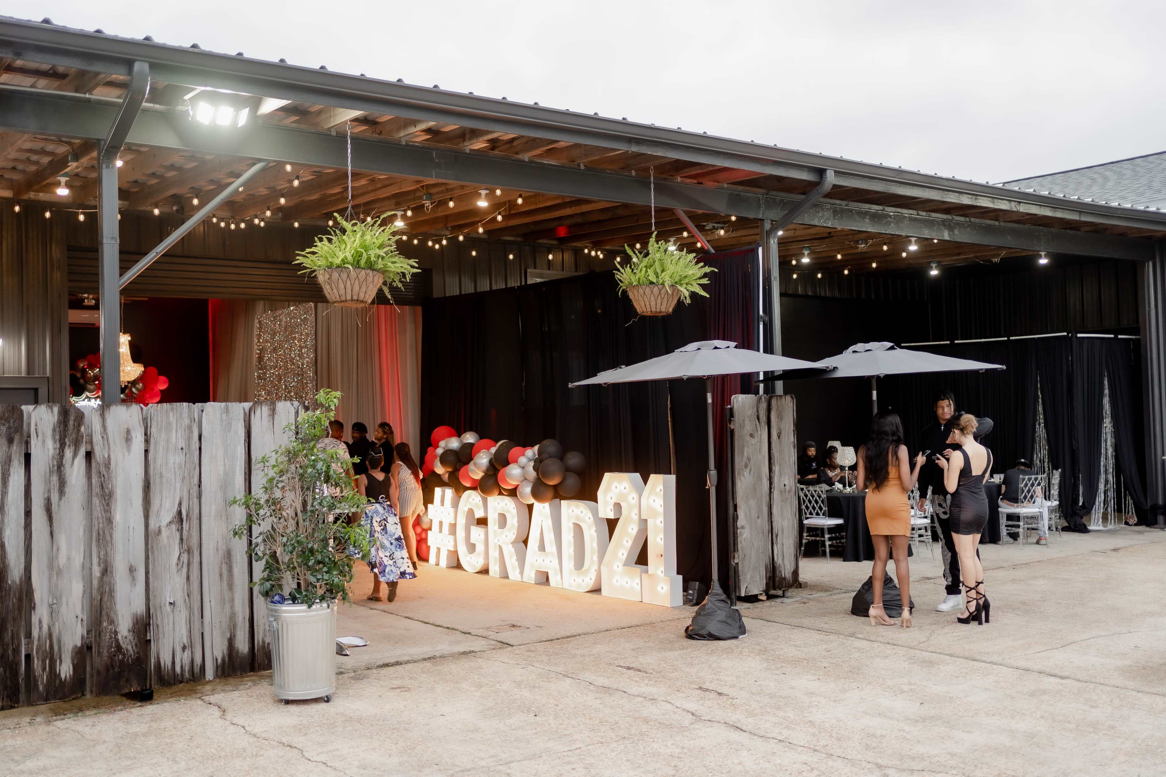 A graduation celebration venue features "#GRAD21" illuminated signage, hanging plants, and guests mingling under umbrellas.