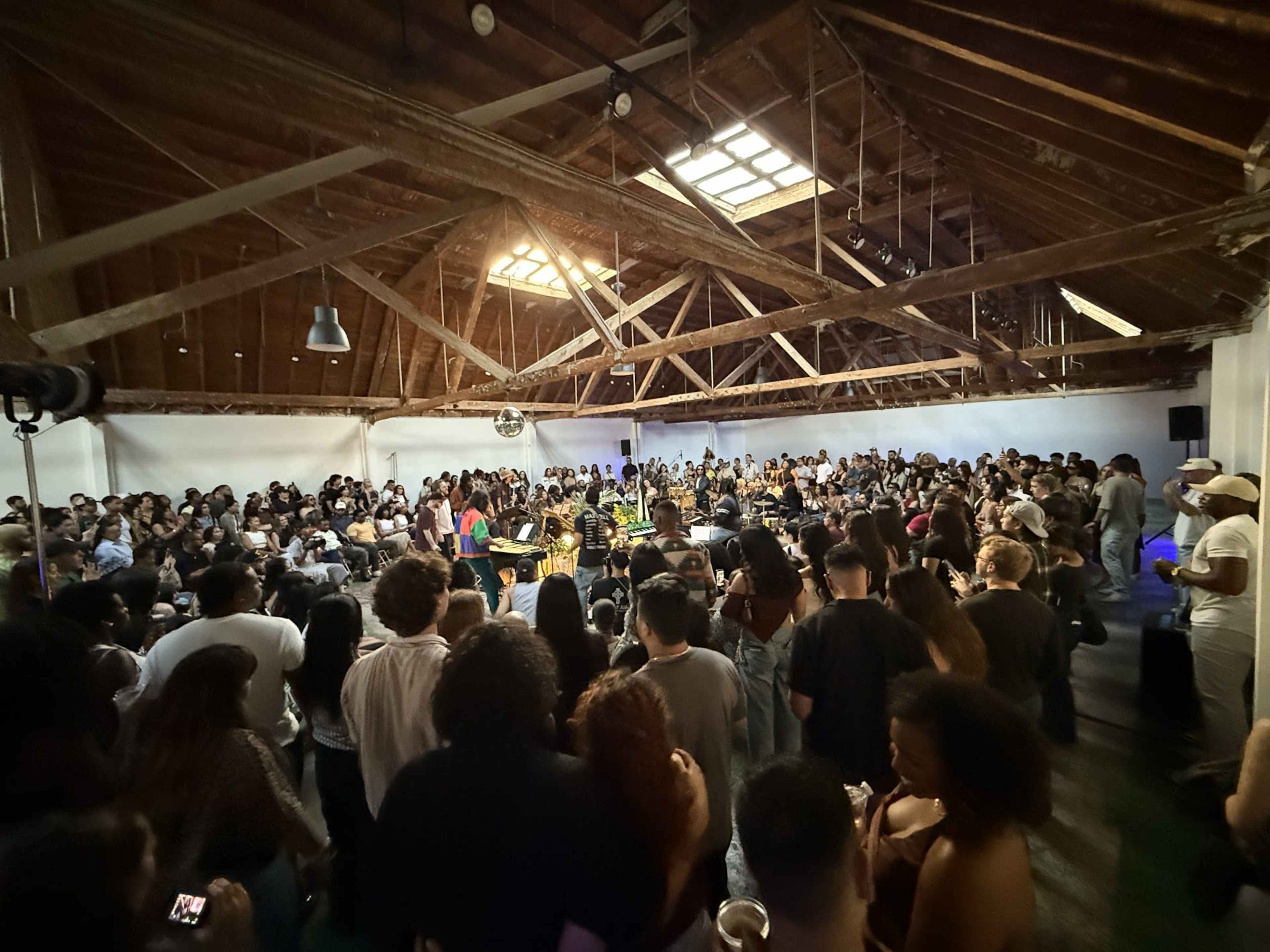 A large crowd of people gathers in an industrial space with wooden beams, watching a performance at the center.