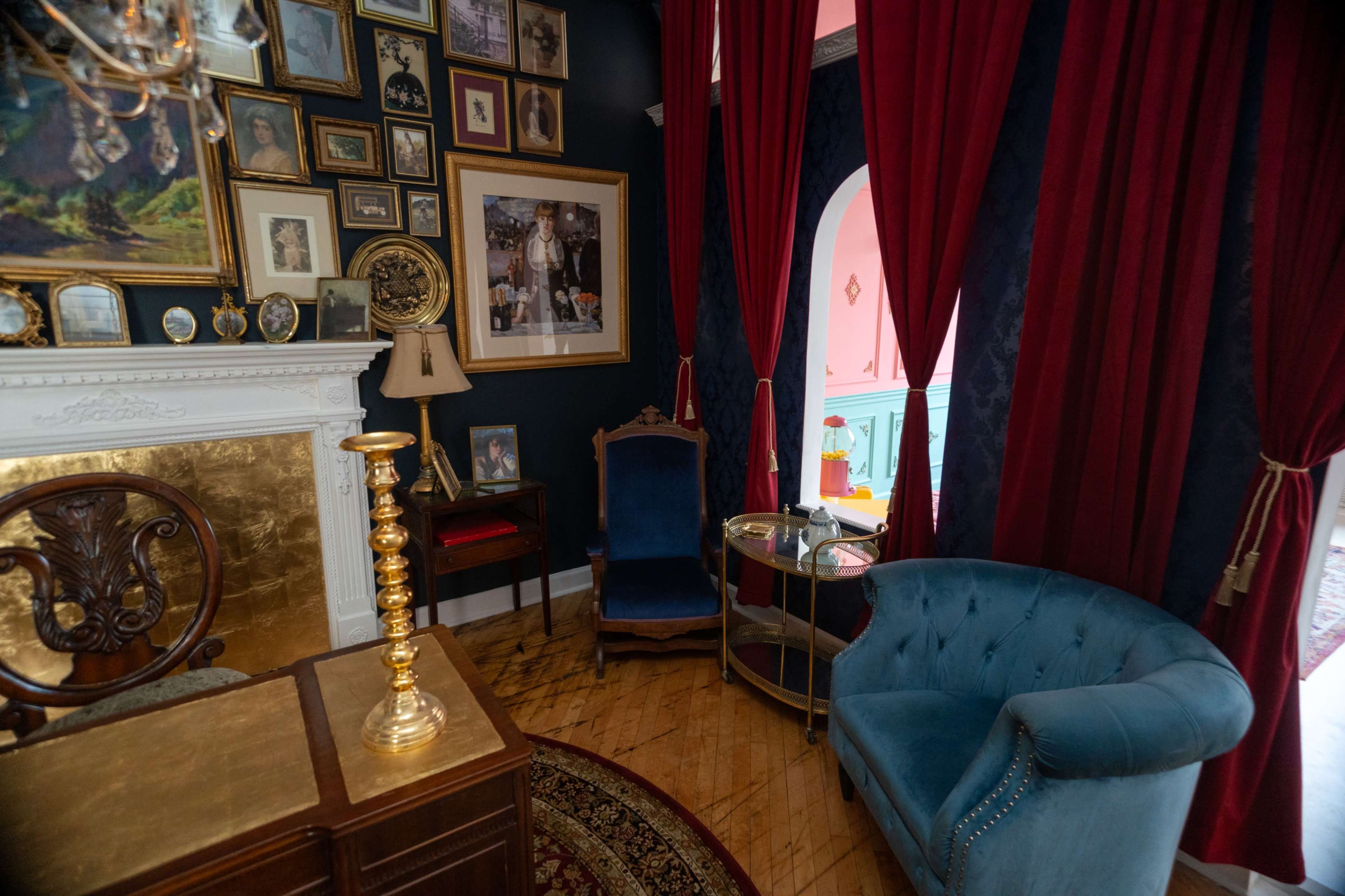 The image depicts a cozy room featuring a gold-accented fireplace, a blue velvet armchair, a series of framed pictures on the dark walls, and red curtains framing an arched doorway.