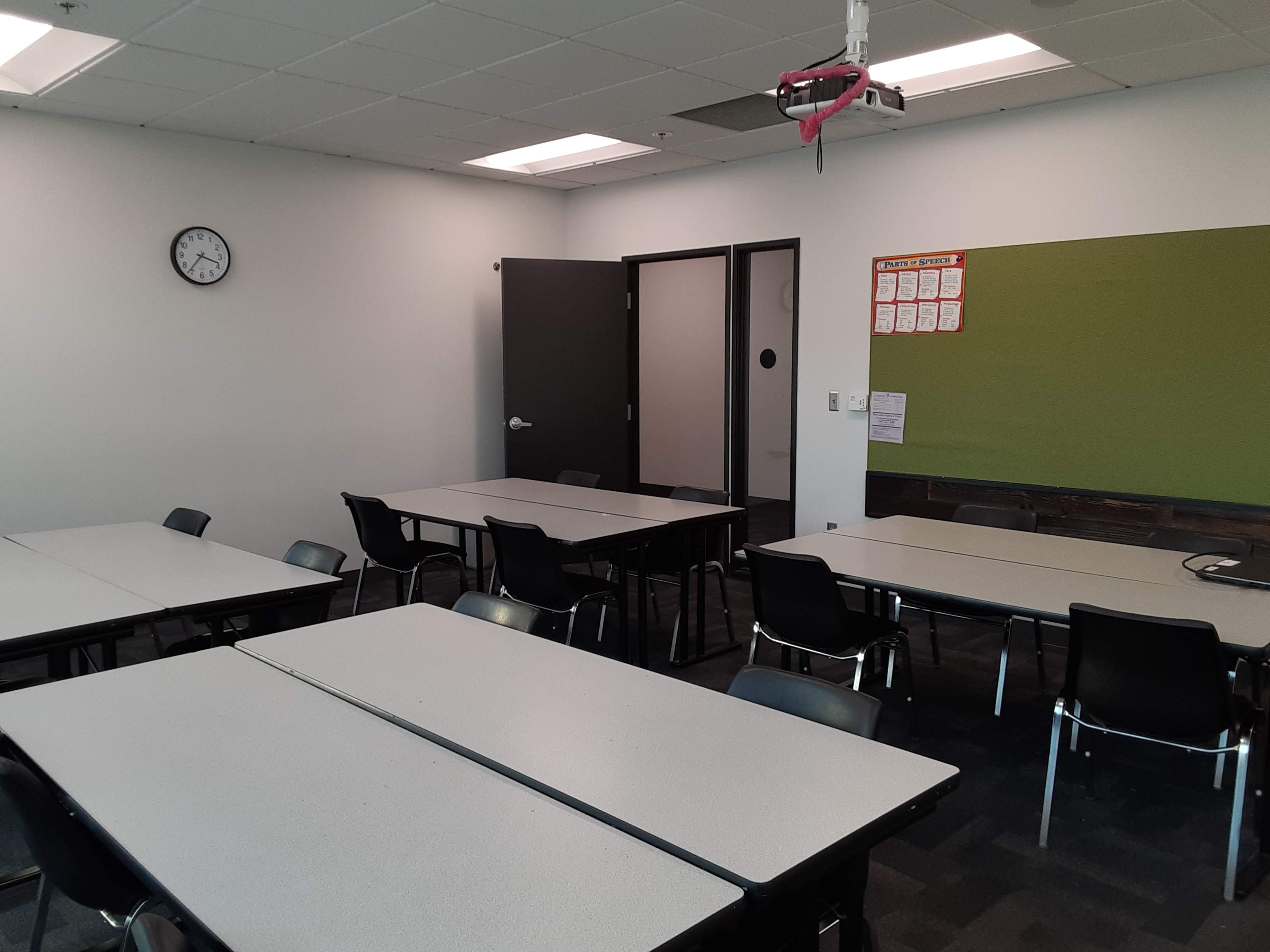 A classroom features several tables arranged with chairs, a clock on the wall, a green board, and a door leading to another room.
