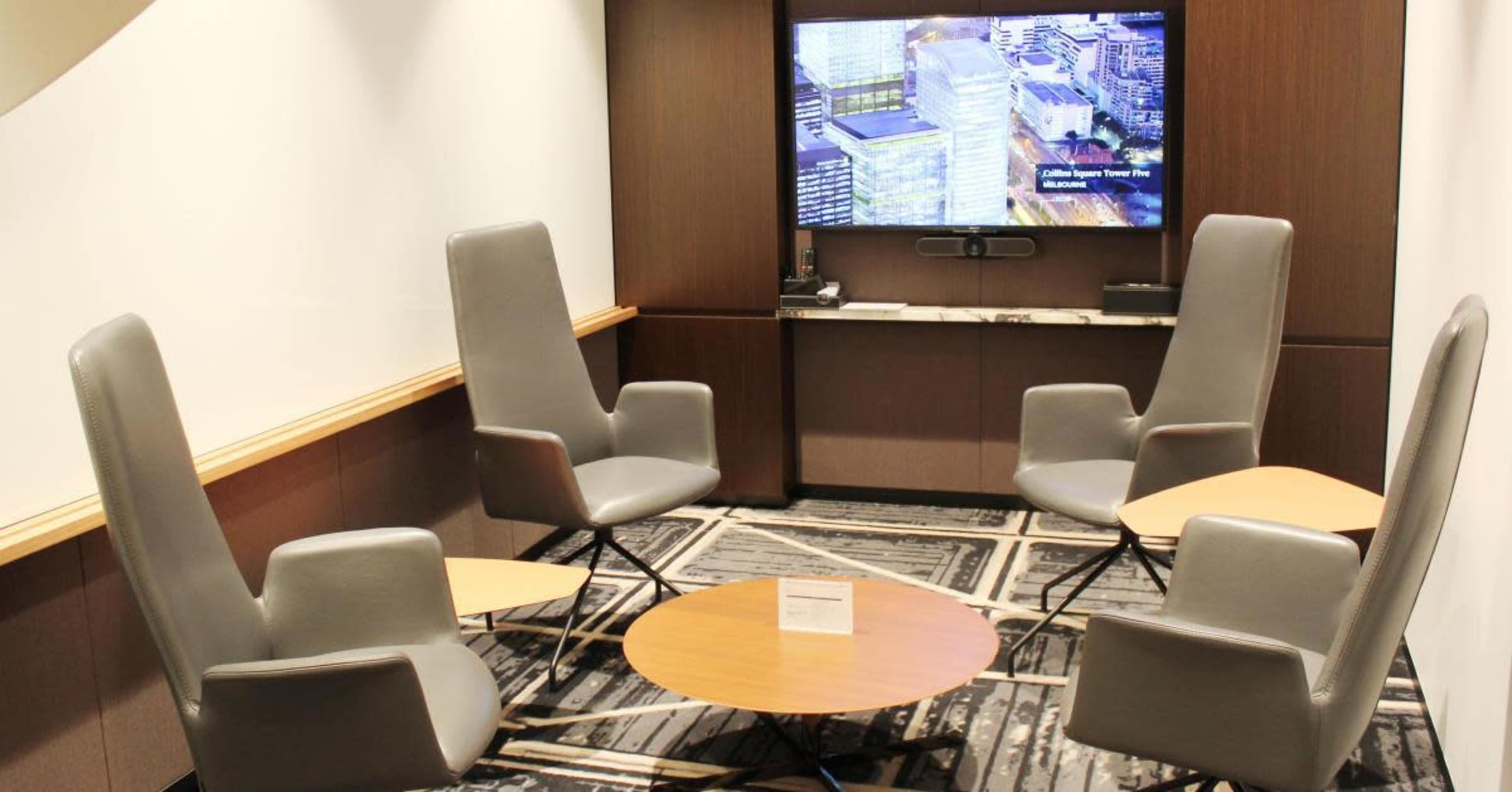 The image shows a modern seating area with four gray high-back chairs arranged around a circular wooden table, accompanied by a large screen displaying a building image on the wall.