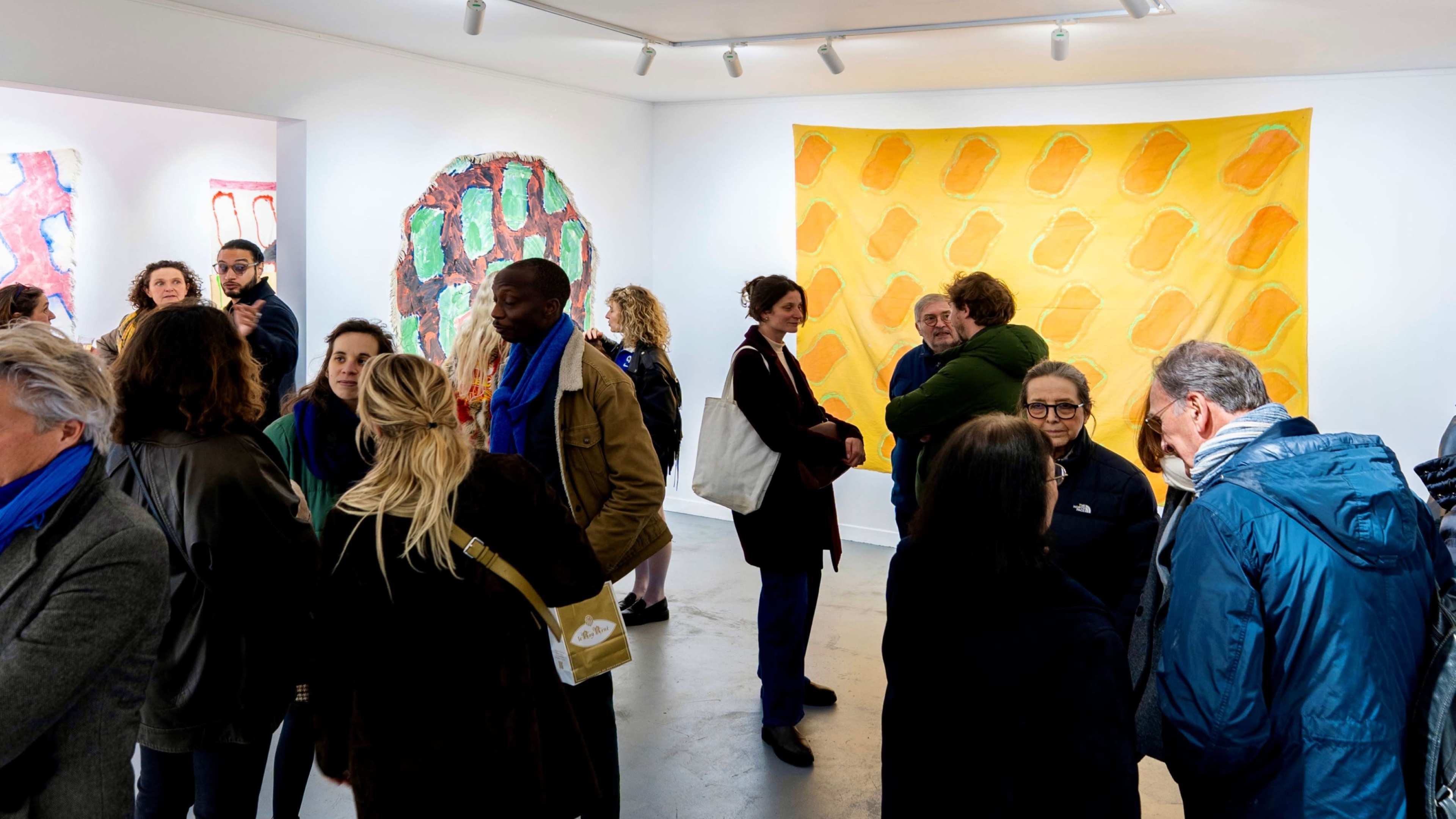 A diverse group of people gathers in an art gallery, engaging in conversations while various colorful artworks adorn the walls.