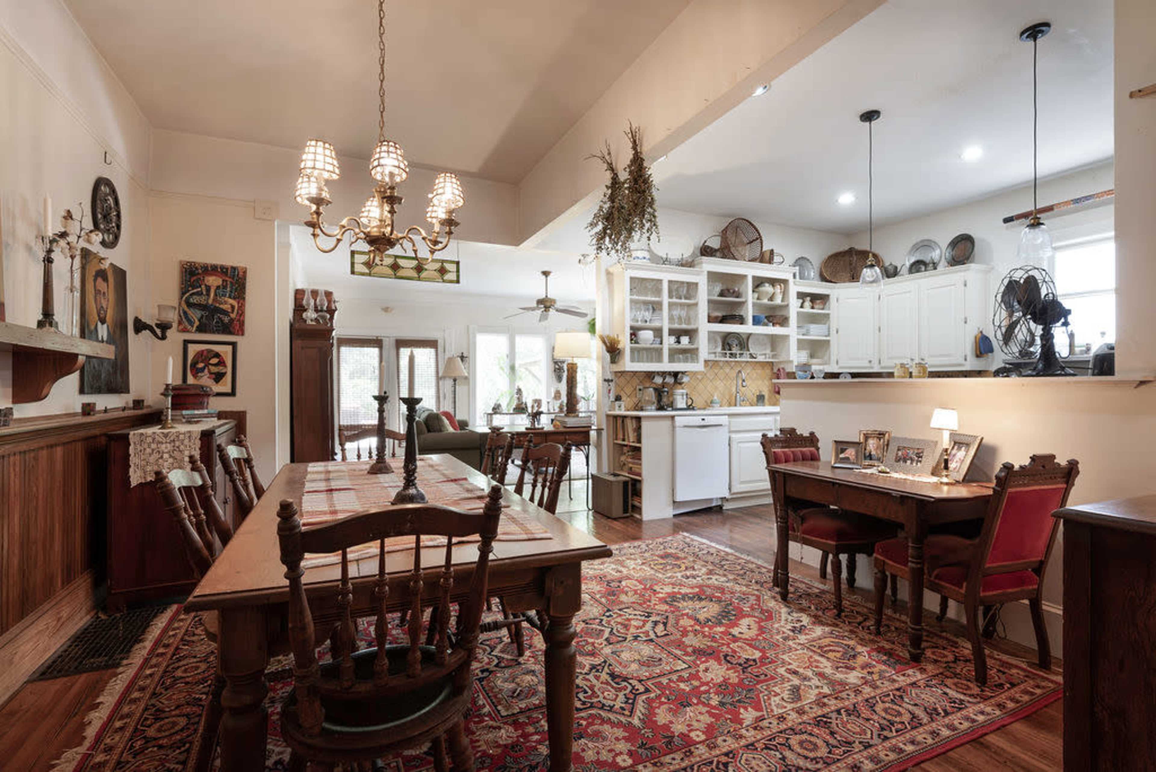 The image shows a spacious dining area with wooden furniture, a large rug, and an open kitchen featuring white cabinetry and appliances.