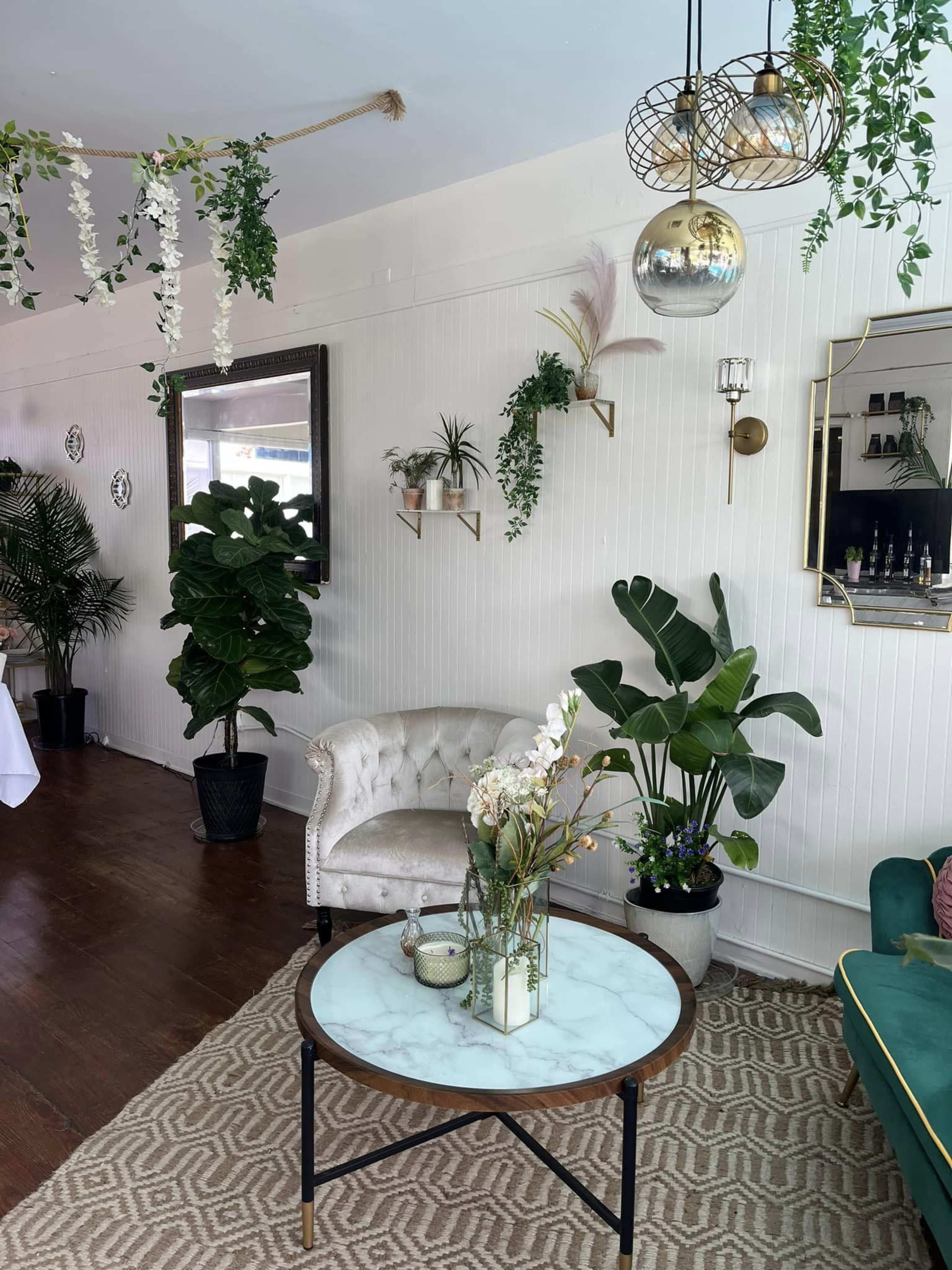 The image depicts a stylish interior space featuring a white wall, a plush armchair, a round table with a marble top, decorative plants, and elegant lighting fixtures.