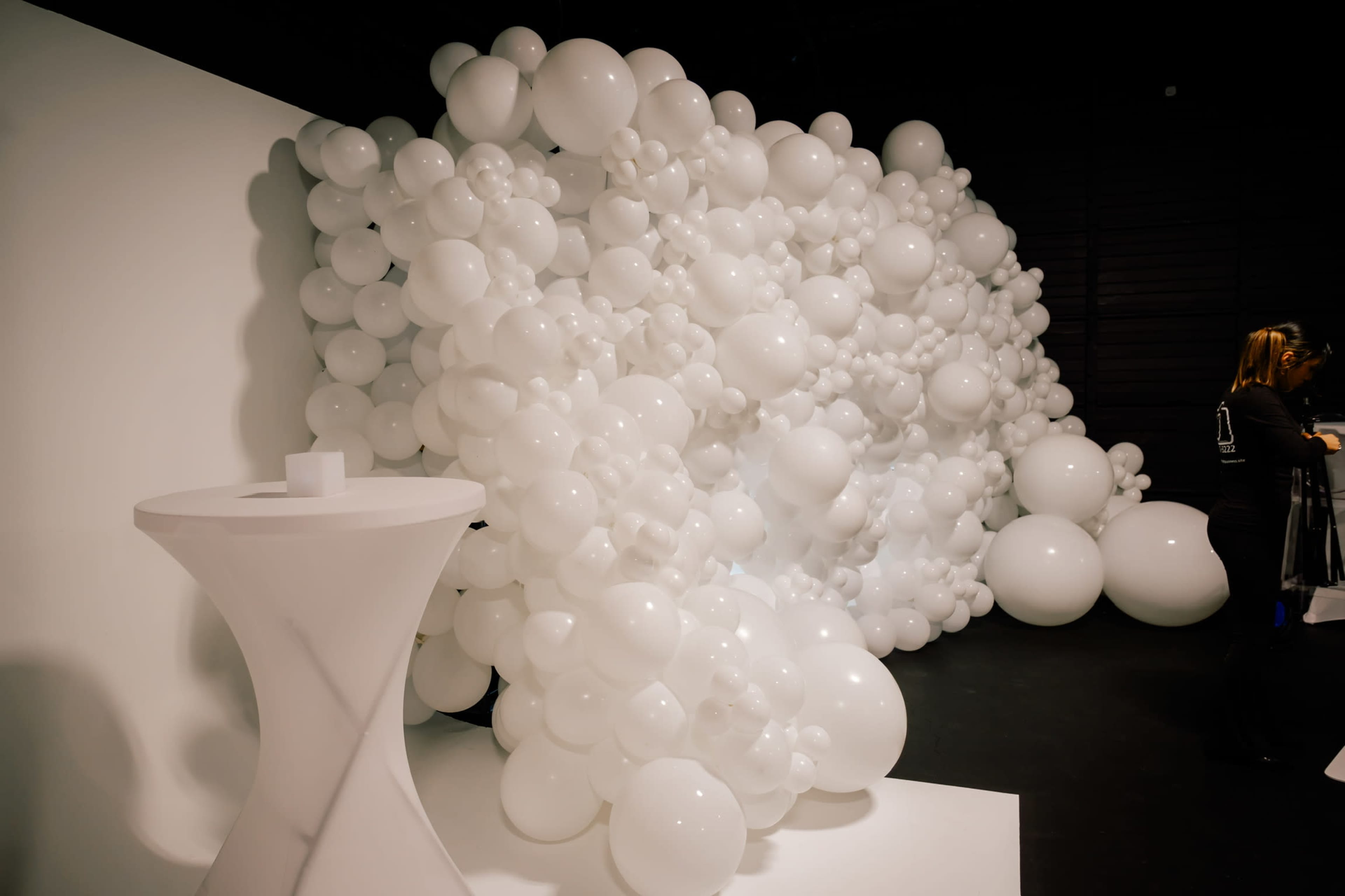 A wall covered with clusters of white balloons is displayed next to a white pedestal table.