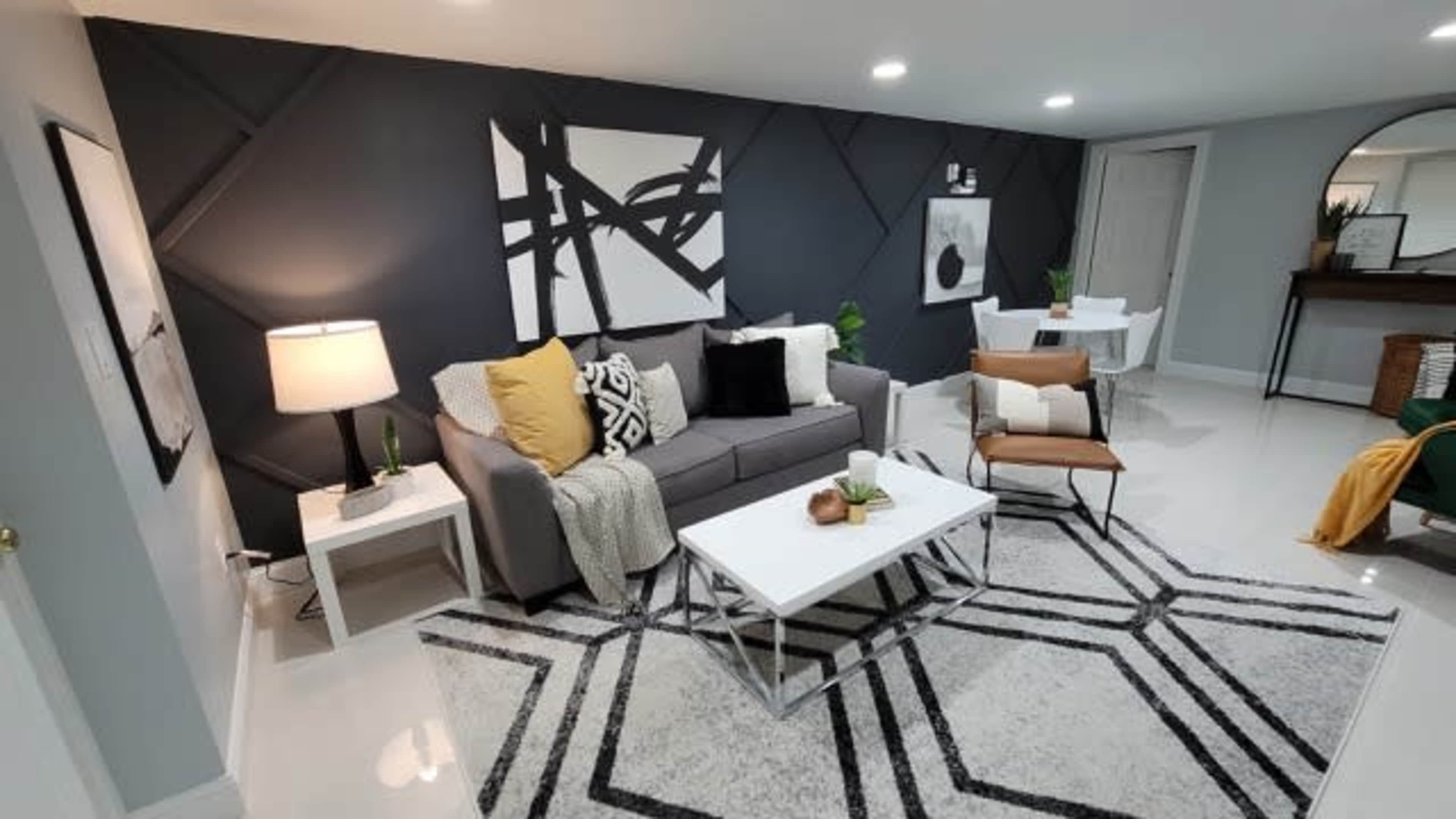 A modern living room features a gray sectional sofa, a geometric rug, a coffee table, and minimalist decor against a dark accent wall.