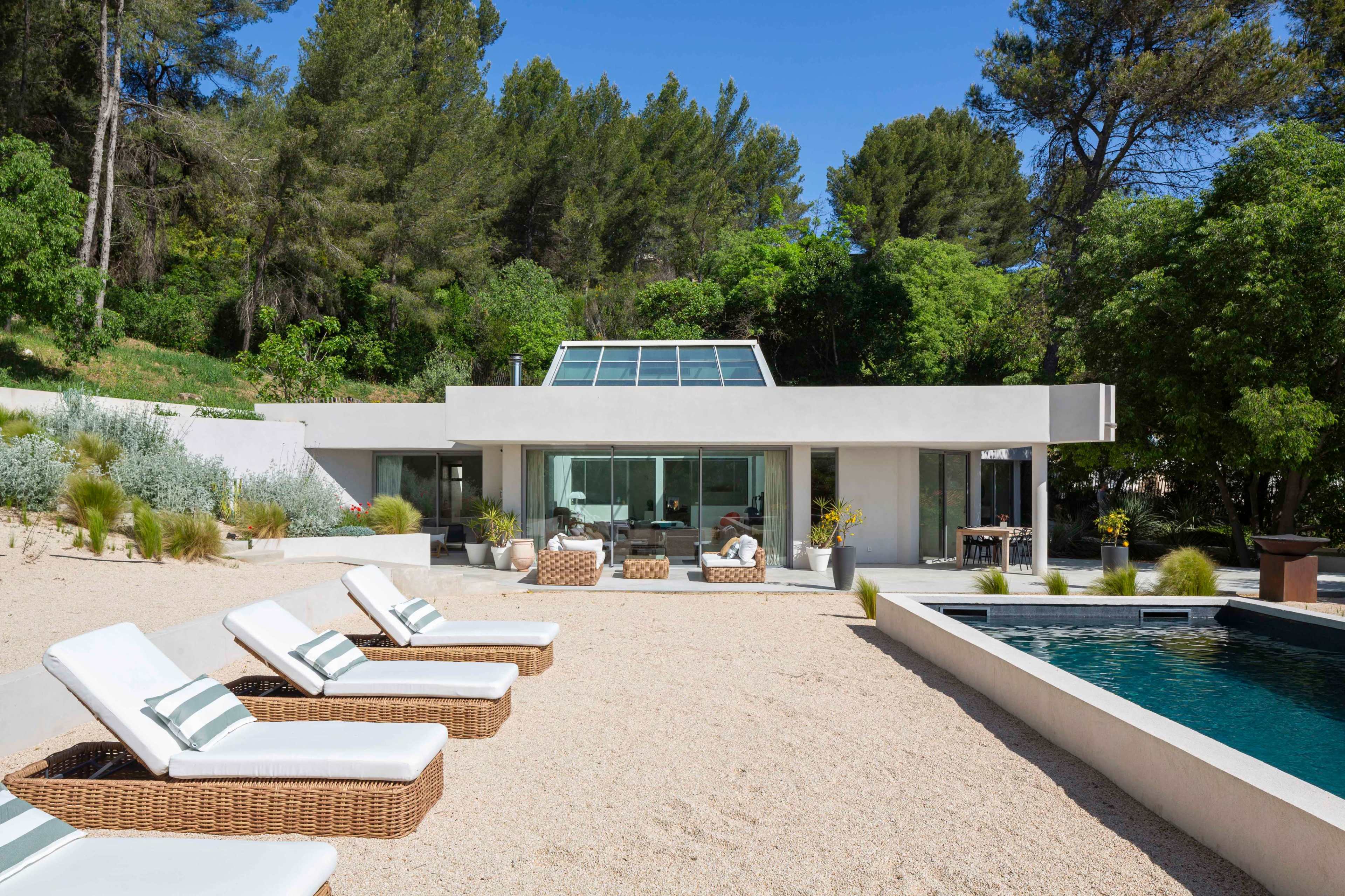 A modern house with large windows and a slanted glass roof is surrounded by a landscaped area featuring lounge chairs and a swimming pool.