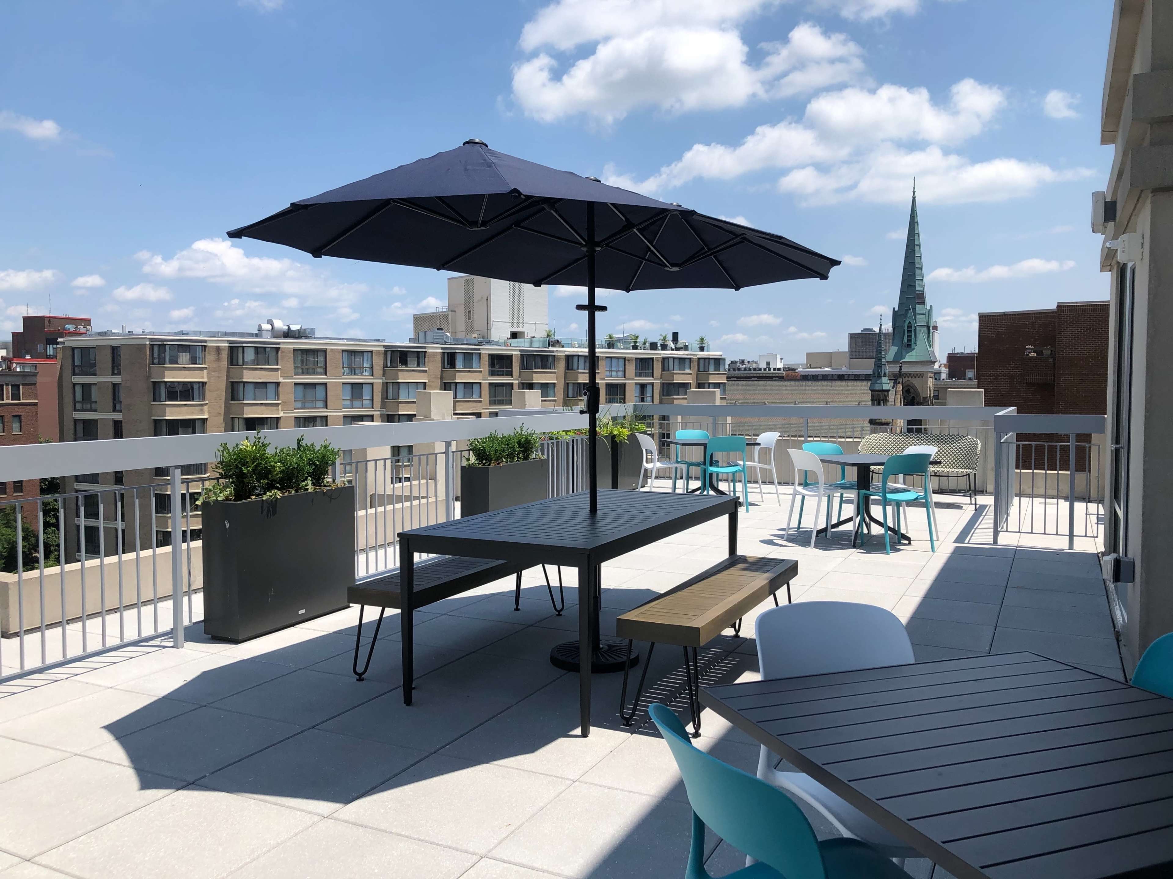 A rooftop terrace with tables, chairs, and a large umbrella, overlooking a cityscape featuring buildings and a steeple in the background.