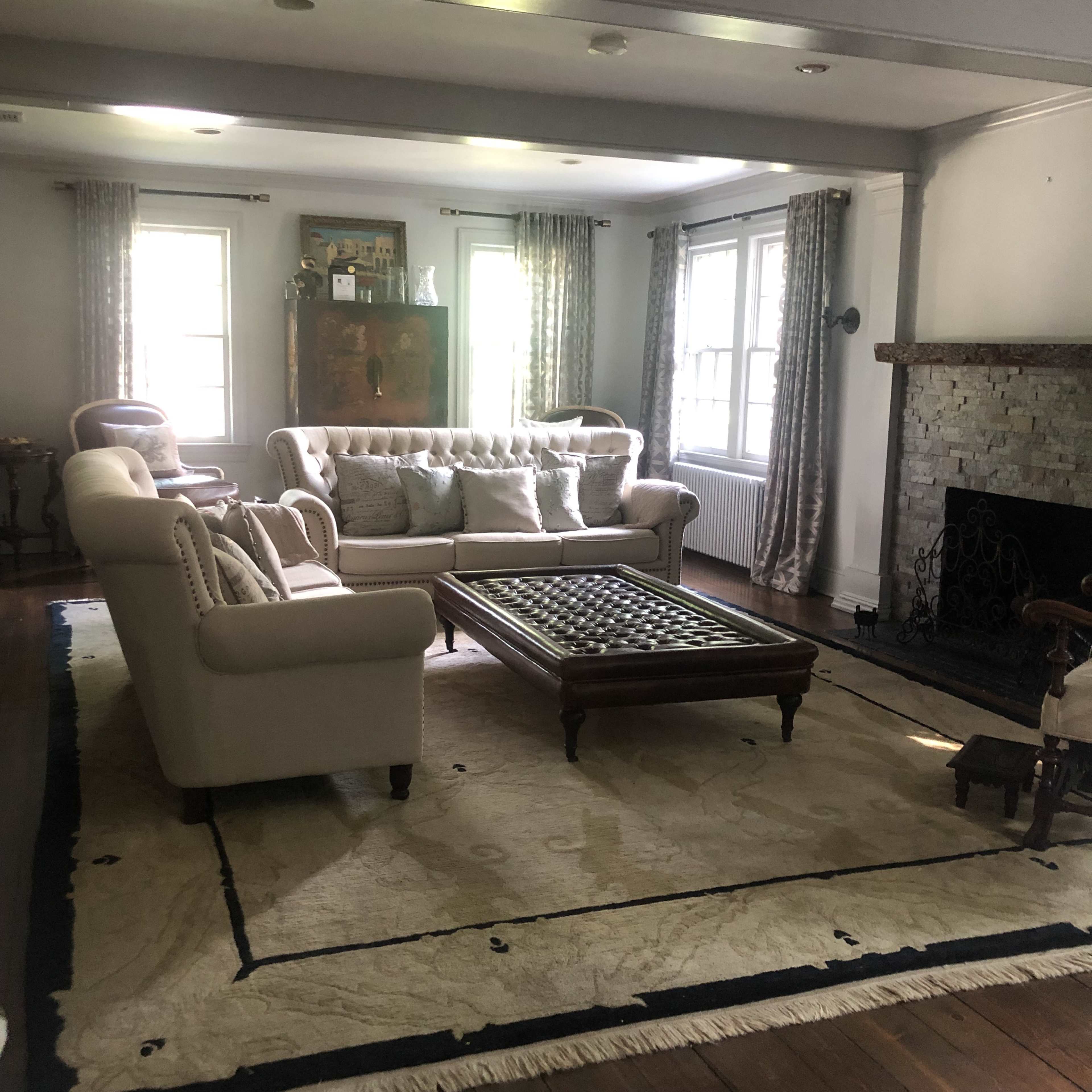 A living room with a white sofa, a tufted coffee table, and a stone fireplace, illuminated by natural light from two windows.