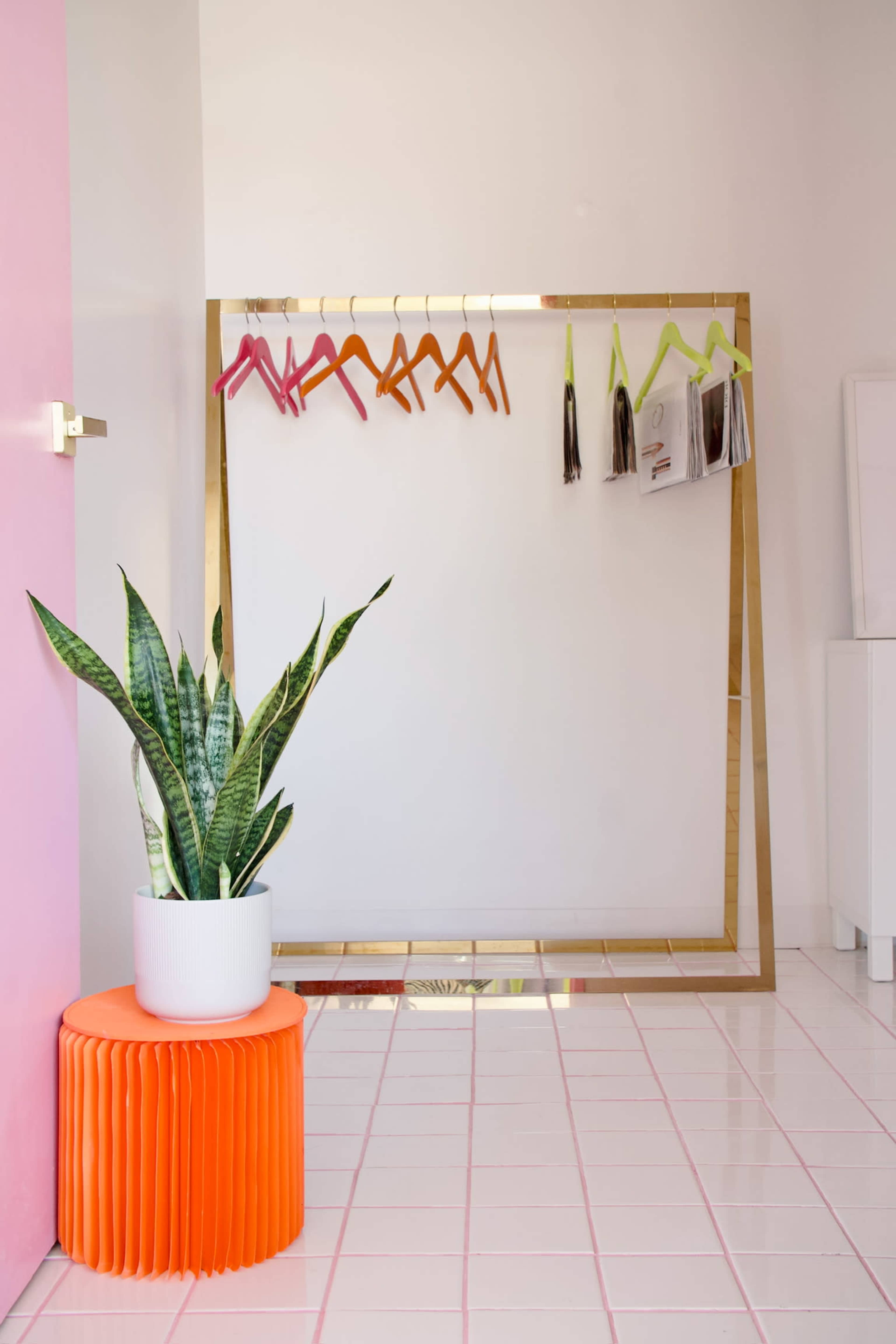 The image shows a minimalist dressing area with a gold-framed mirror, colorful hangers on a rack, a potted plant, and an orange stool.