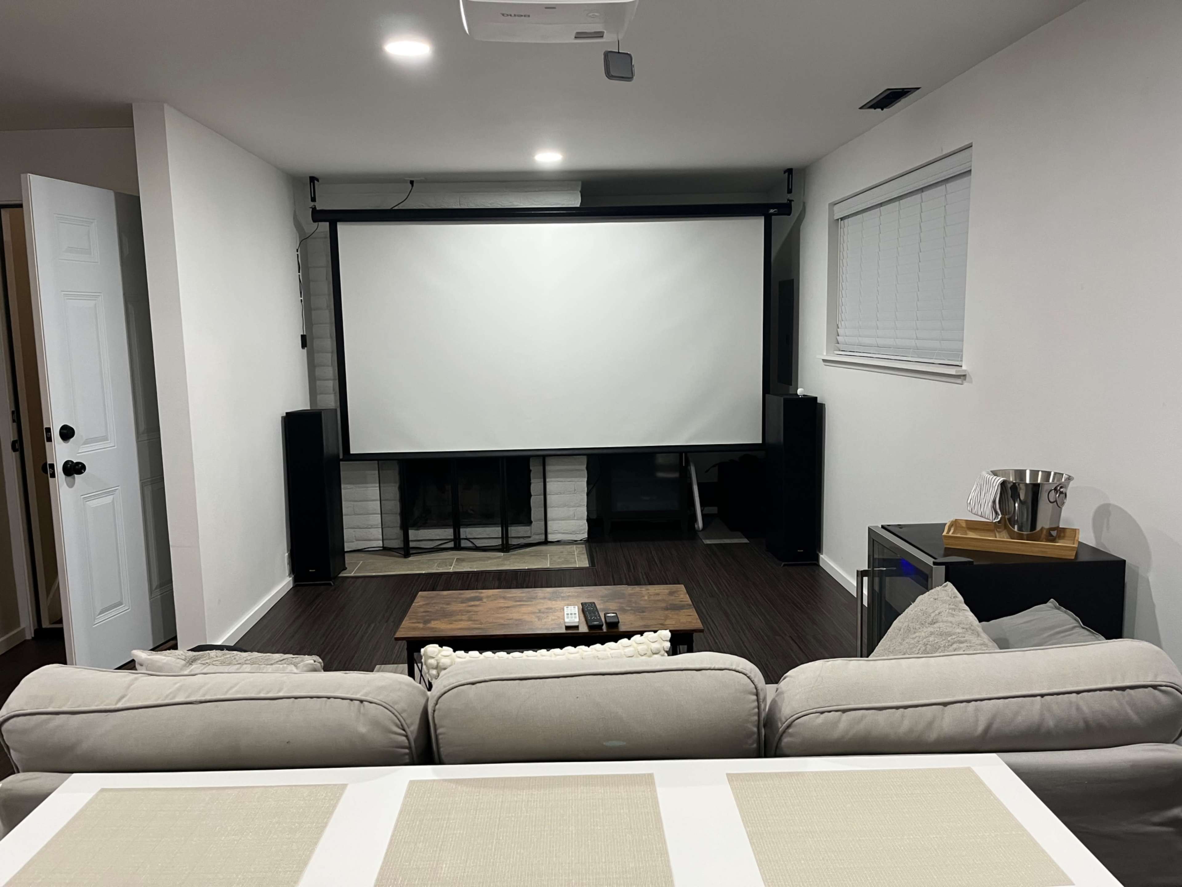 A spacious home theater setup features a large projector screen mounted on the wall, flanked by two speakers, with a comfortable sofa and a coffee table in front.