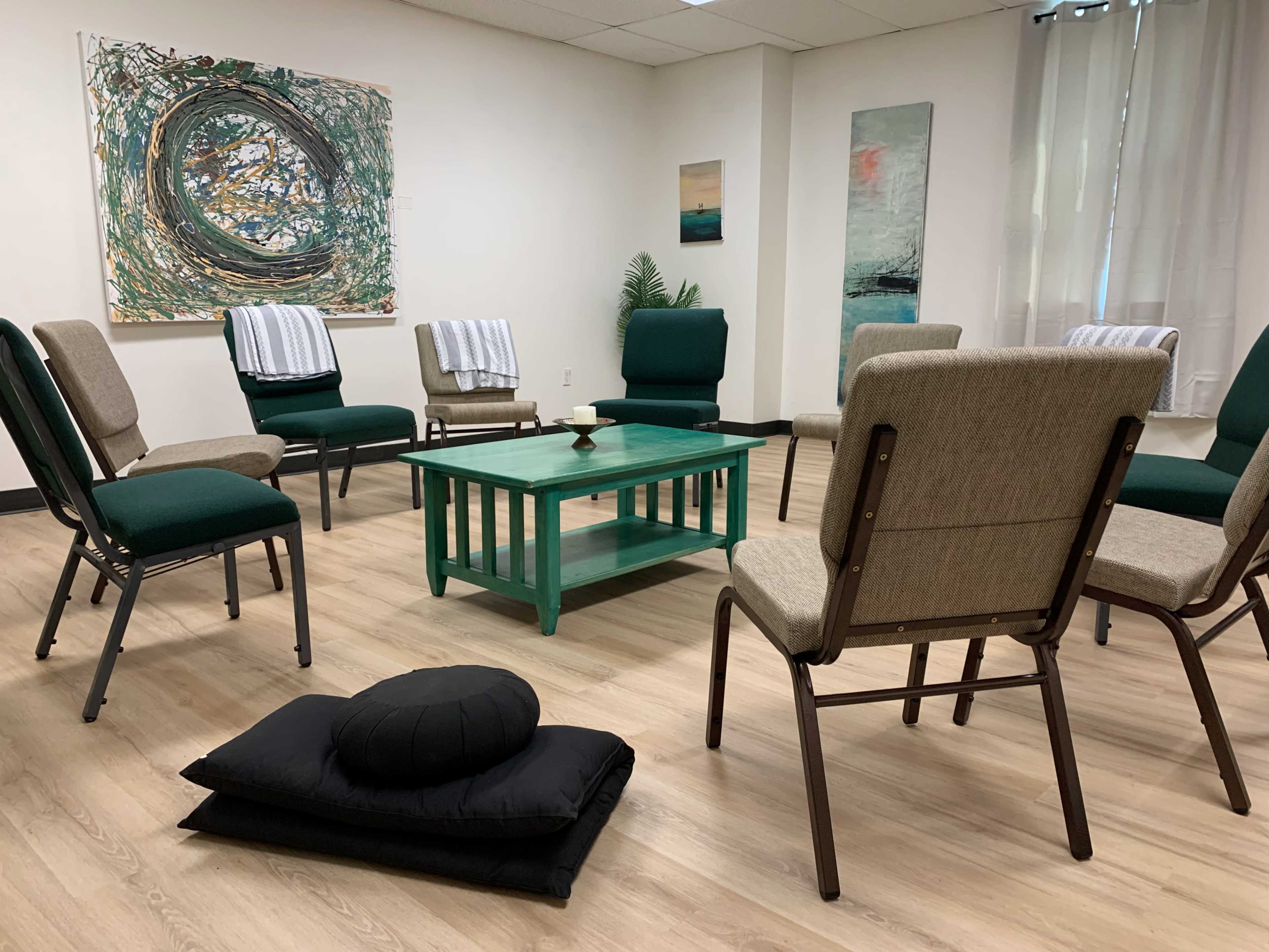 A room with a circular arrangement of chairs around a green coffee table, featuring art on the walls and a black cushion on the floor.