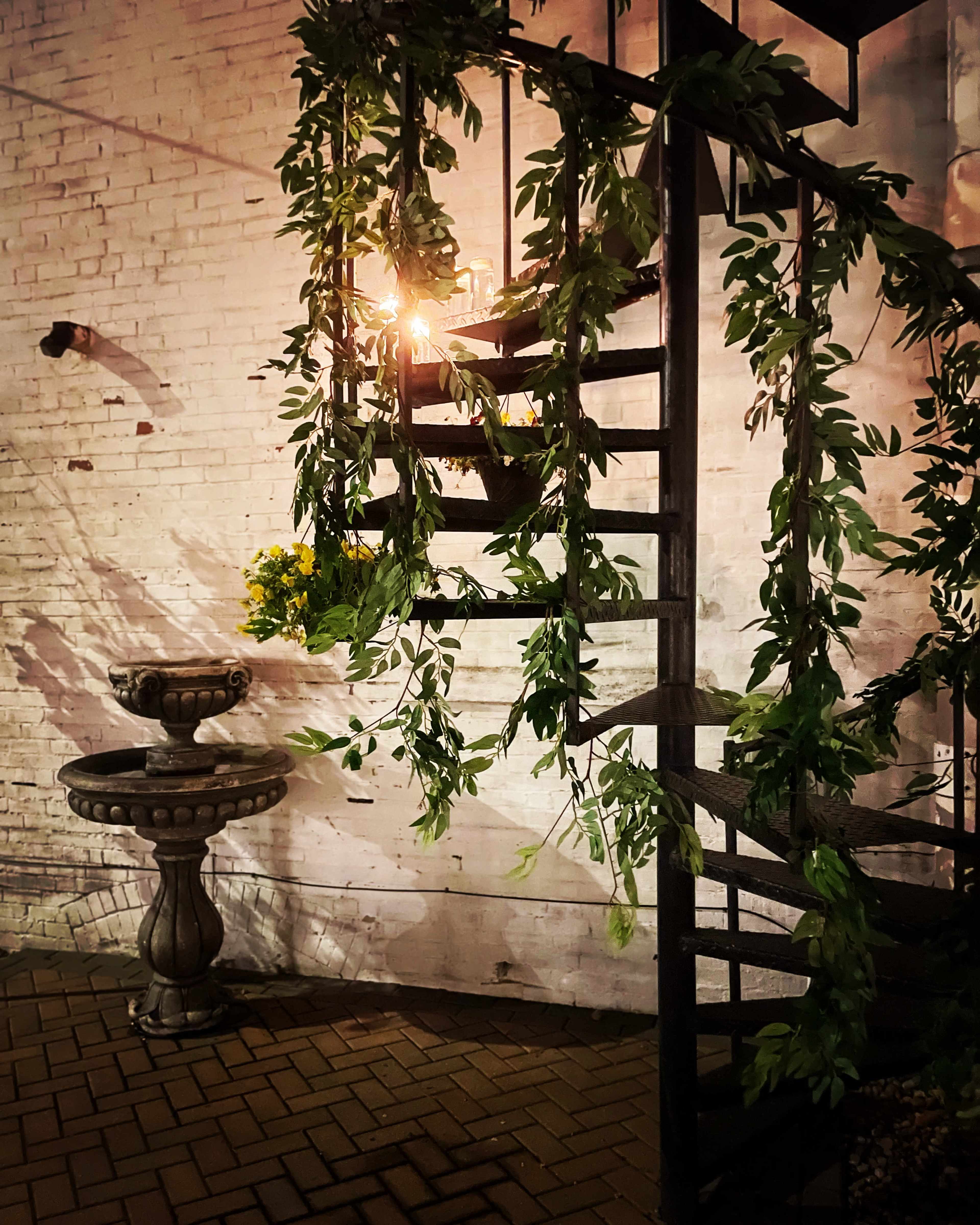 A spiral staircase draped with greenery stands next to a decorative stone fountain illuminated by overhead light.
